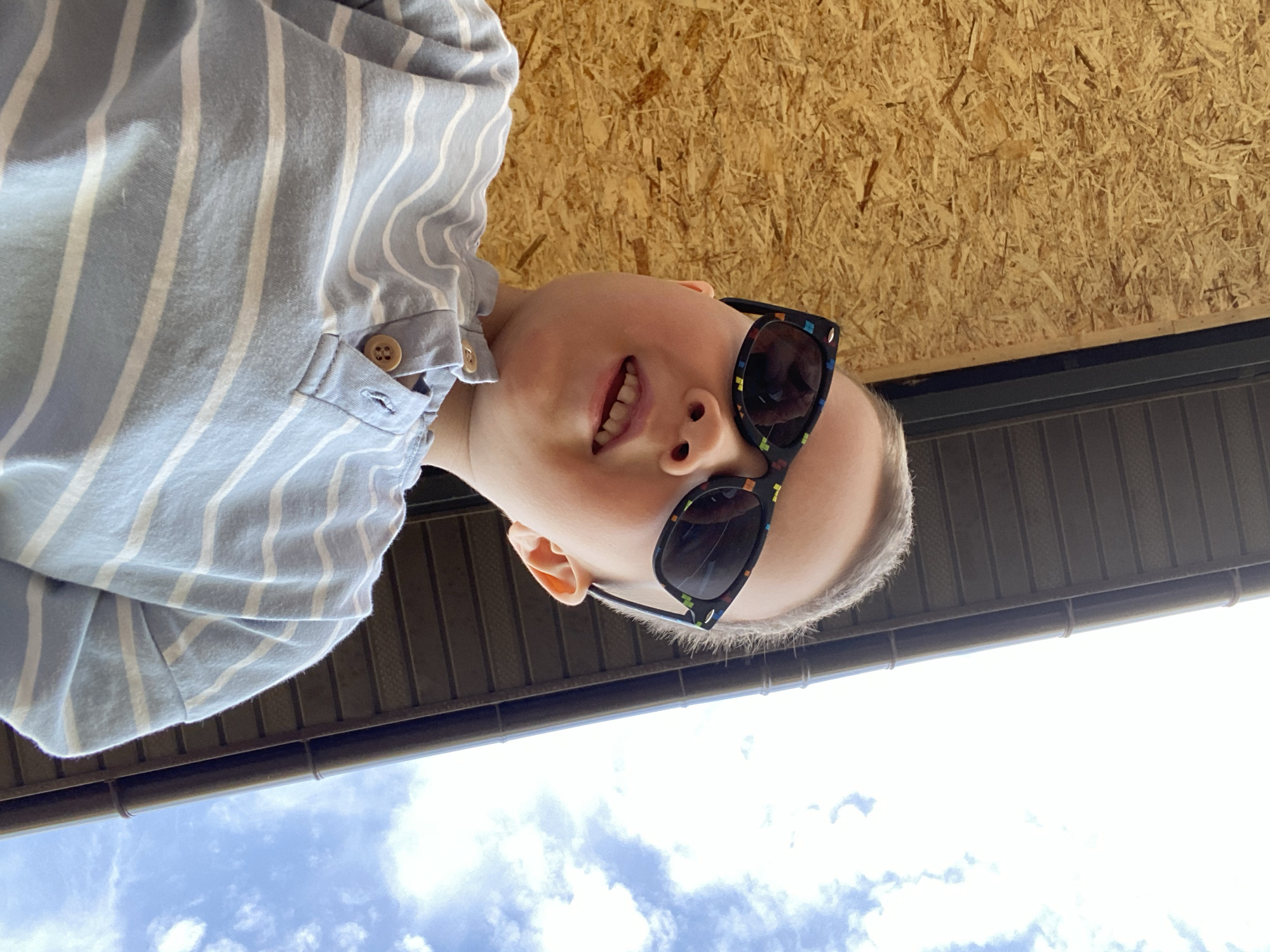 A child wearing sunglasses and a blue and white striped shirt looks down at the camera. The background consists of an unfinished wooden ceiling, a metal overhang, and a blue sky with white clouds. The location cannot be determined as Chaikovsky, Russia, based on the visual data.