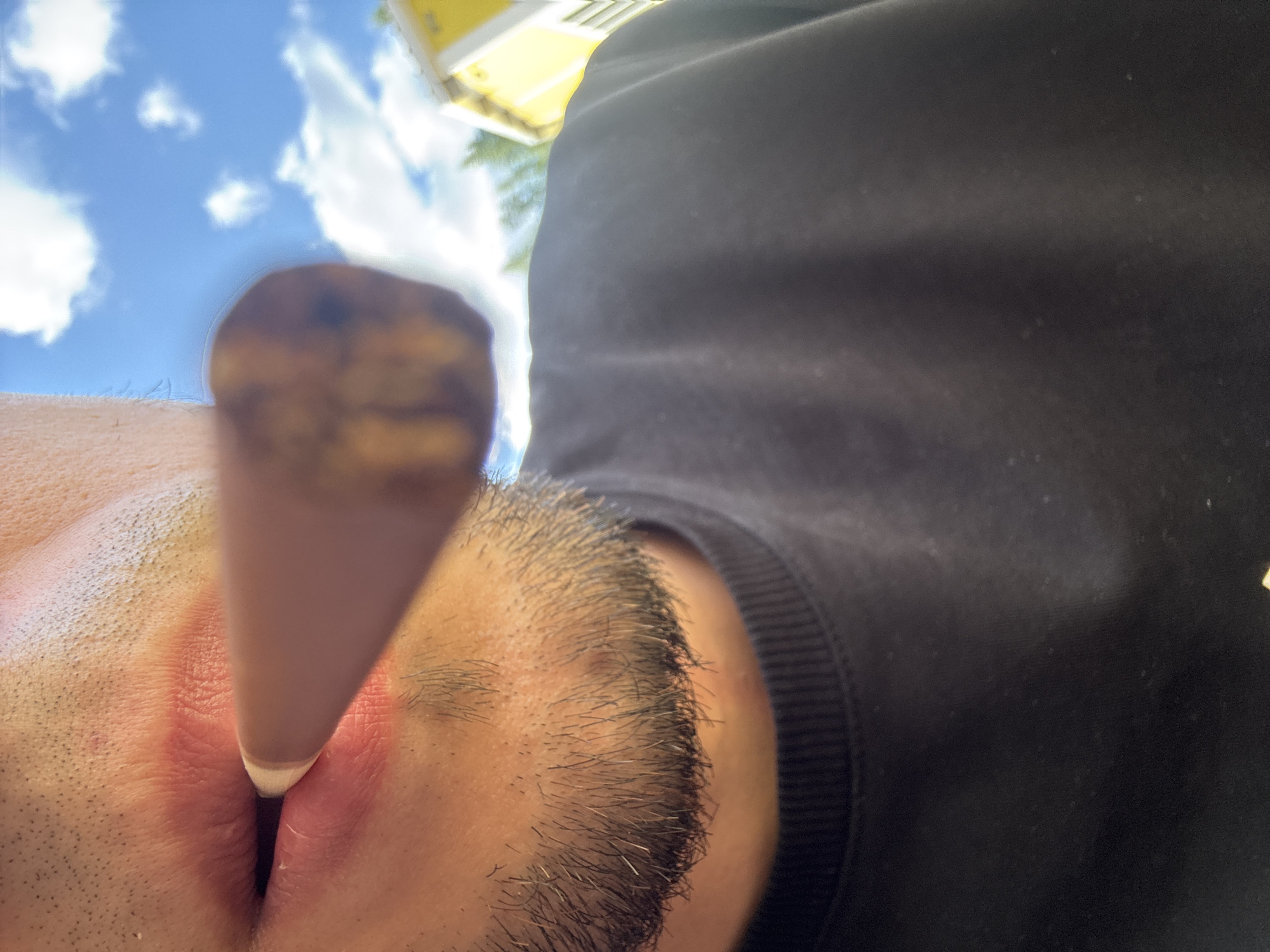 The image is a close-up, low-angle shot of a person's face, with the camera positioned near their nose. The person is wearing a black hoodie, visible on the right side of the frame. A light-colored object, possibly a stylus or pen, is positioned in front of the person's nose and mouth area, with its tip slightly between the lips. The background shows a blue sky with white clouds and a hint of a yellow building or structure. The overall lighting suggests it is daytime. The composition creates a somewhat abstract perspective of the human face and the surrounding environment. There is no visible text.