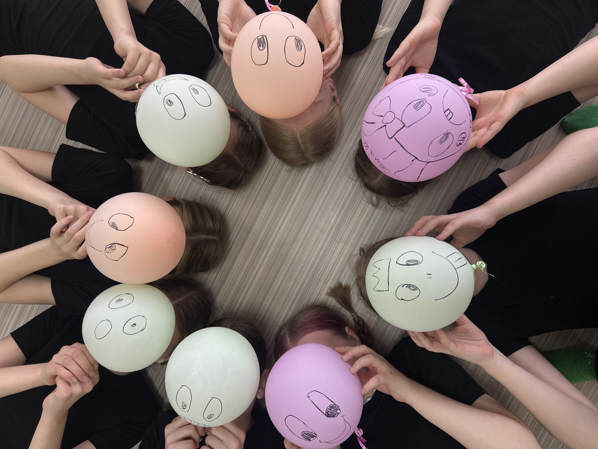 Eight individuals, likely children or youths, are lying on their backs on a light-colored wooden floor in a circular formation, with their heads positioned towards the center. Dressed uniformly in black clothing, each person holds a pastel-colored balloon (shades of light green/white, pink, and peach) directly over their face, creating the playful illusion that the balloons are their heads. Each balloon features a unique, whimsical face drawn with black marker, depicting various eye shapes, eyebrows, and expressions. Small ribbons or ties are attached to some balloons.
On one pink balloon with a large bow design, the names "Саша" (Sasha) and "Катя" (Katya) are written vertically. Another pink balloon, featuring an open-mouthed face drawing, has the word "Сама." (Sama) written on its side. The image is captured from an overhead perspective, highlighting the creative and joyful activity. This group interaction takes place indoors, likely during a game or photoshoot, in Votkinsk, Russia.
