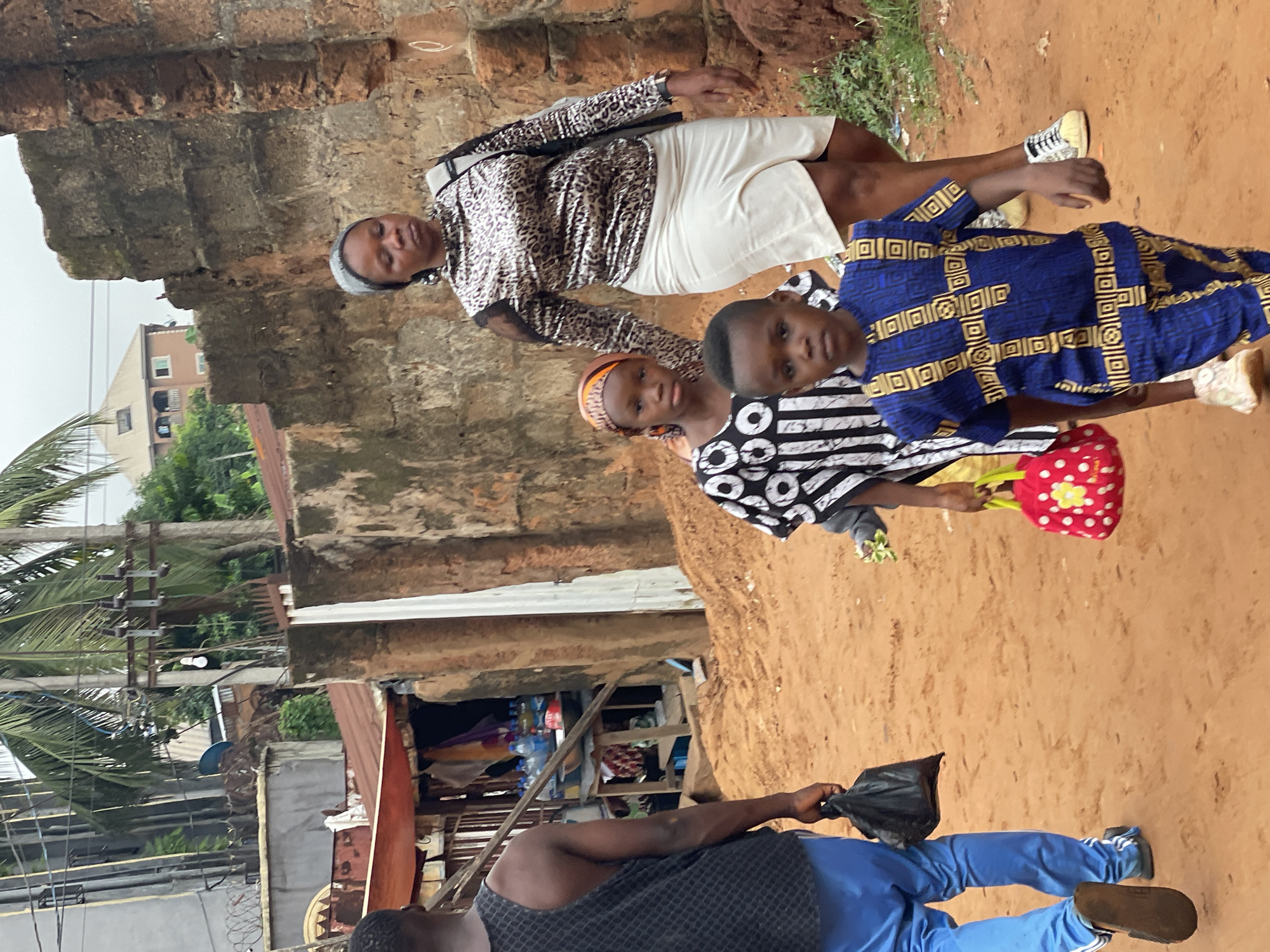 The image shows a group of three individuals standing on a dirt ground adjacent to a weathered brick wall and a corrugated metal structure in Nkpor, Nigeria. An adult female is wearing a leopard-print top and white shorts. To her right are two children, one wearing a blue and gold patterned garment and the other in a black and white striped shirt, holding a red polka-dot bag. In the foreground, a man is partially visible, wearing blue trousers and a black tank top, holding a black plastic bag. The background features trees, power lines, and buildings typical of an urban environment.