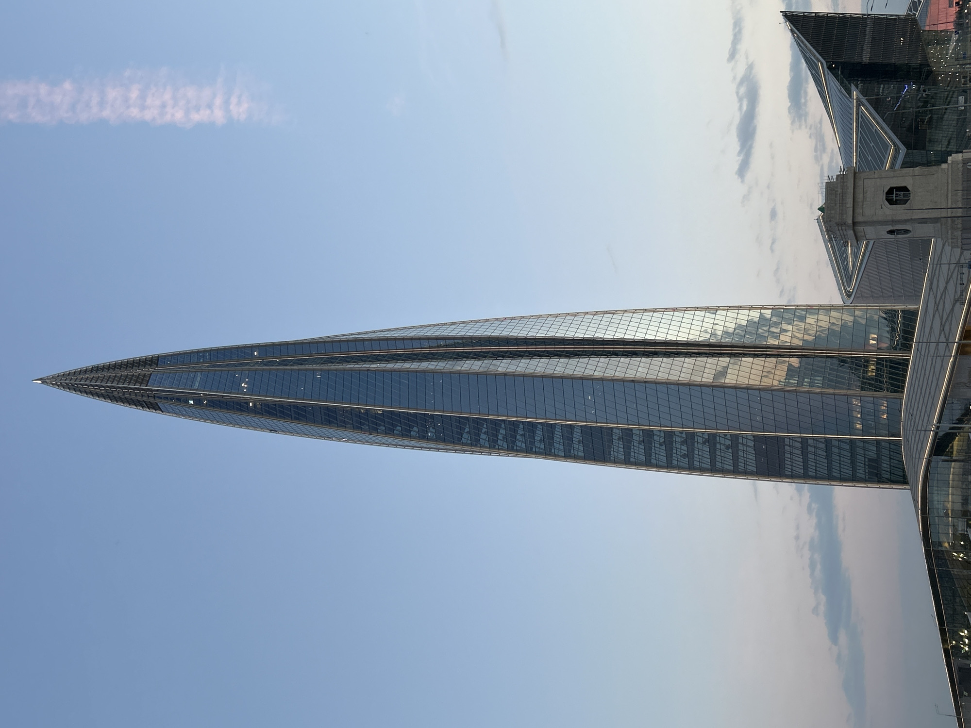 The image shows the Lakhta Center in Saint Petersburg, Russia. The skyscraper is angled across the frame, its spire pointing towards the left. The building is composed of multiple glass panels, reflecting the sky and surrounding environment. The sky is mostly clear with some clouds near the top right corner. Another building, possibly part of the same complex, is partially visible in the upper right of the frame. There is a vapor trail in the sky towards the left side of the image.