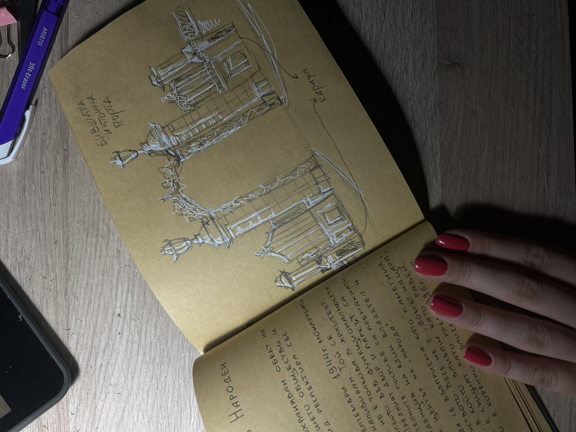 The image shows an open notebook on a wooden surface. The right page features a white ink sketch of an ornate gate, with text written above it in the same ink, reading "БИВШАТА ИЗТОЧНА ПОРТА" (Former East Gate) and "КАРАУЛ" (Guardhouse) with an arrow pointing to a section of the gate. The left page contains handwritten text in Bulgarian, which appears to be a historical description. The text discusses a protected object, mentioning its cultural significance and "respectful architecture." It also refers to events in September 1944 and the monarchy, as well as its function as a functional house and park. Further text details its transformation into a "home for the Tsar" and its preservation. The notebook is open, revealing a person's left hand with bright red nail polish, resting on the lower part of the left page. A portion of a dark-colored object, possibly a phone or tablet, is visible in the upper left corner of the image. A purple pen with an "ARISTO" label and "3ff-Eraser" marking lies on the wooden surface, partially obscuring the top of the notebook. The lighting creates shadows, suggesting it is likely indoors or in a shaded outdoor area.