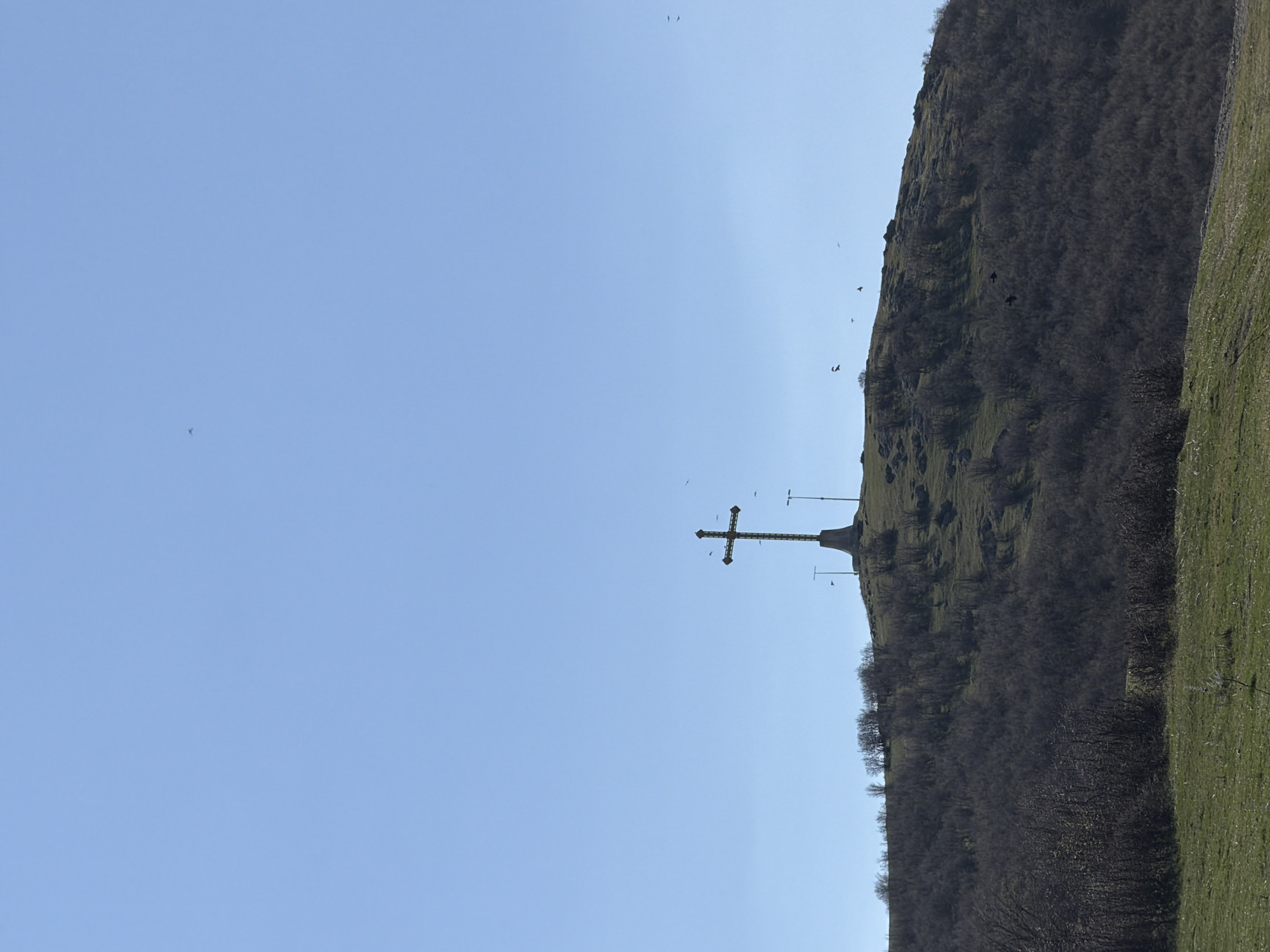 The image shows a large metal cross on a hilltop, presumably in or near Bata, Bulgaria, as the user specified. The cross is dark in color and sits atop a concrete or stone base. To the right of the cross, the hill slopes down, covered in dark, dense vegetation, possibly trees and shrubs. Patches of green grass are visible on the lower right of the frame. Above the hill, a clear blue sky is visible. Several birds are seen flying in the sky. The image appears to be taken during daylight.