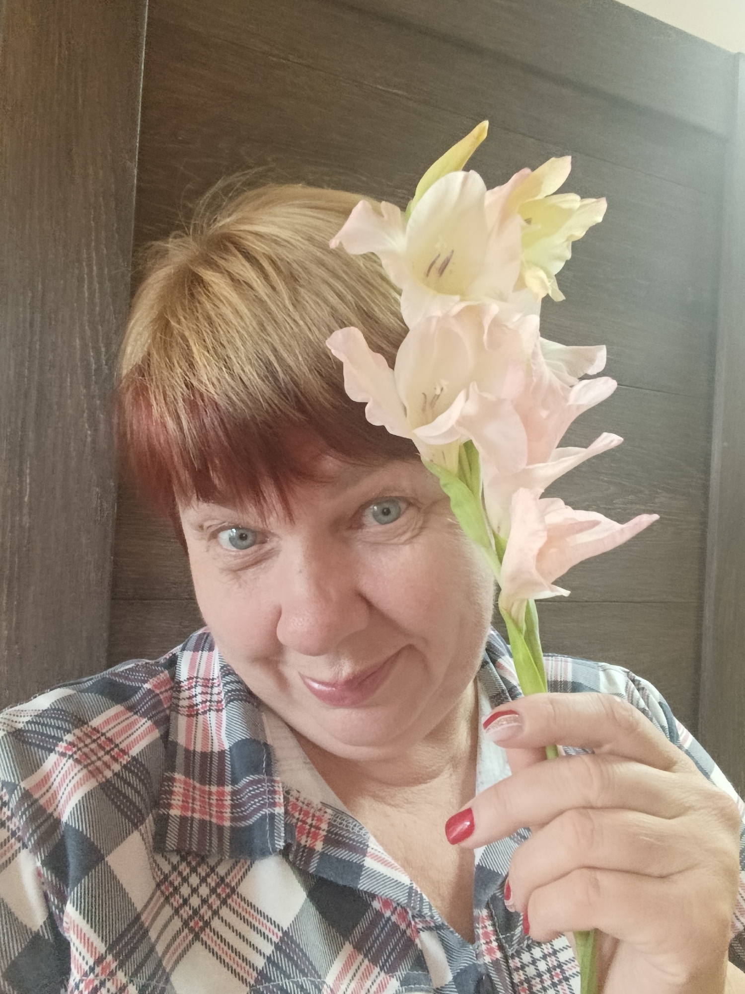 A mid-shot, outdoor portrait shows a middle-aged woman with short, reddish-blonde hair and blue eyes. She is smiling at the camera and holding a gladiolus flower stem with several pale pink and yellow blooms to the right of her head. She is wearing a blue, white, and pink plaid button-down shirt with a white undershirt visible at the collar. Her fingernails are painted red. The background appears to be a wooden wall, with a vertical line suggesting a door or panel. The lighting is soft, with a brighter area to the right indicating a possible light source. The image appears to be taken in Svietlahorsk, Belarus.