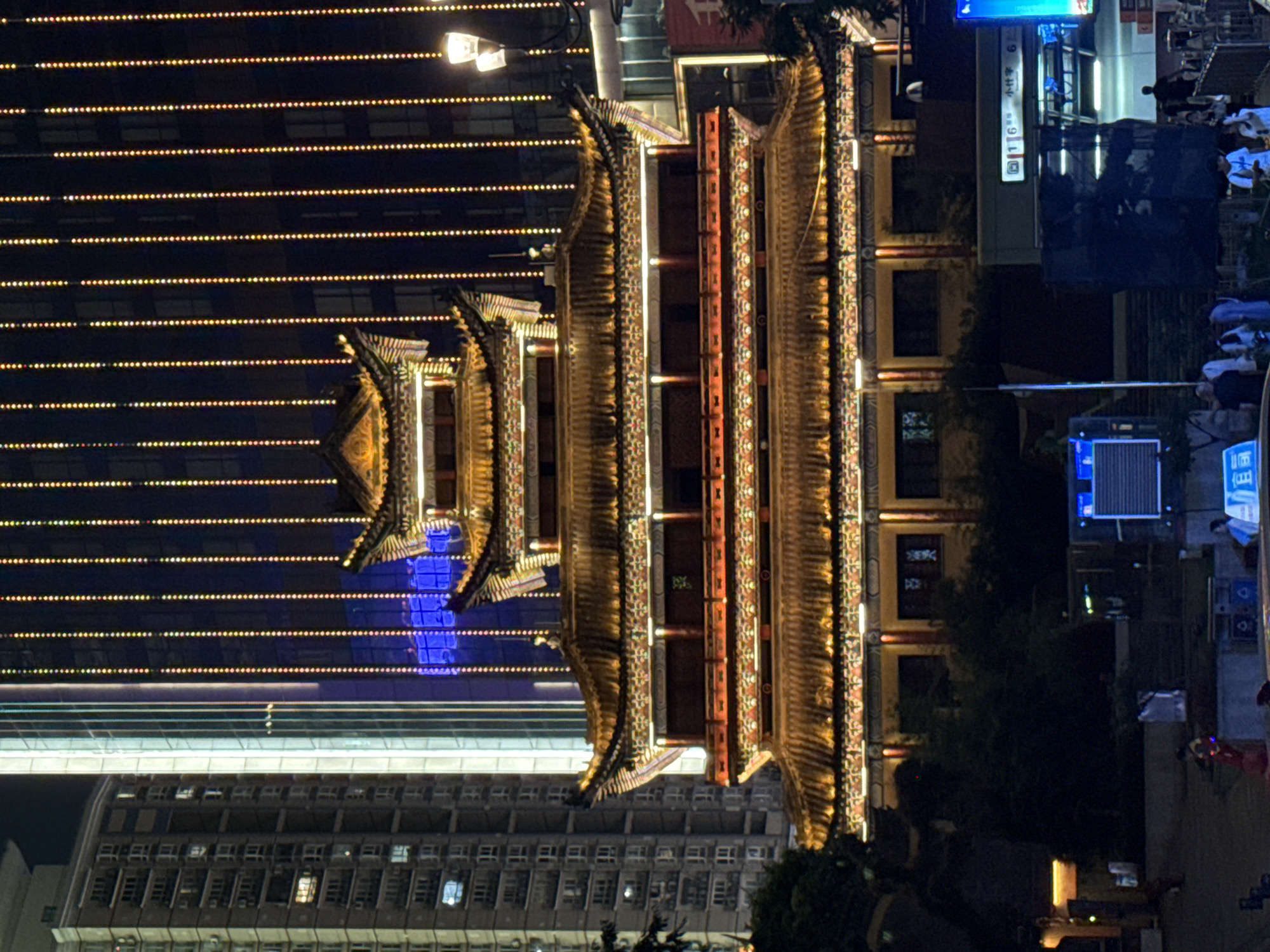The image depicts a night scene in the Yuzhong District of Chongqing, China, featuring a traditional multi-tiered Chinese pavilion illuminated with warm lights, set against a backdrop of modern high-rise buildings. The pavilion's ornate, tiered rooflines are outlined in yellow light, with red accents on the lower levels. Vertical strings of colorful lights, predominantly yellow and red, cascade down the darker facade of the buildings behind the pavilion, creating a vibrant visual contrast. In the foreground, the scene transitions to a street level with a few people visible, and a glimpse of what appears to be a subway station entrance with signage. The overall atmosphere is one of urban dynamism, blending historical architectural elements with contemporary city life at night.