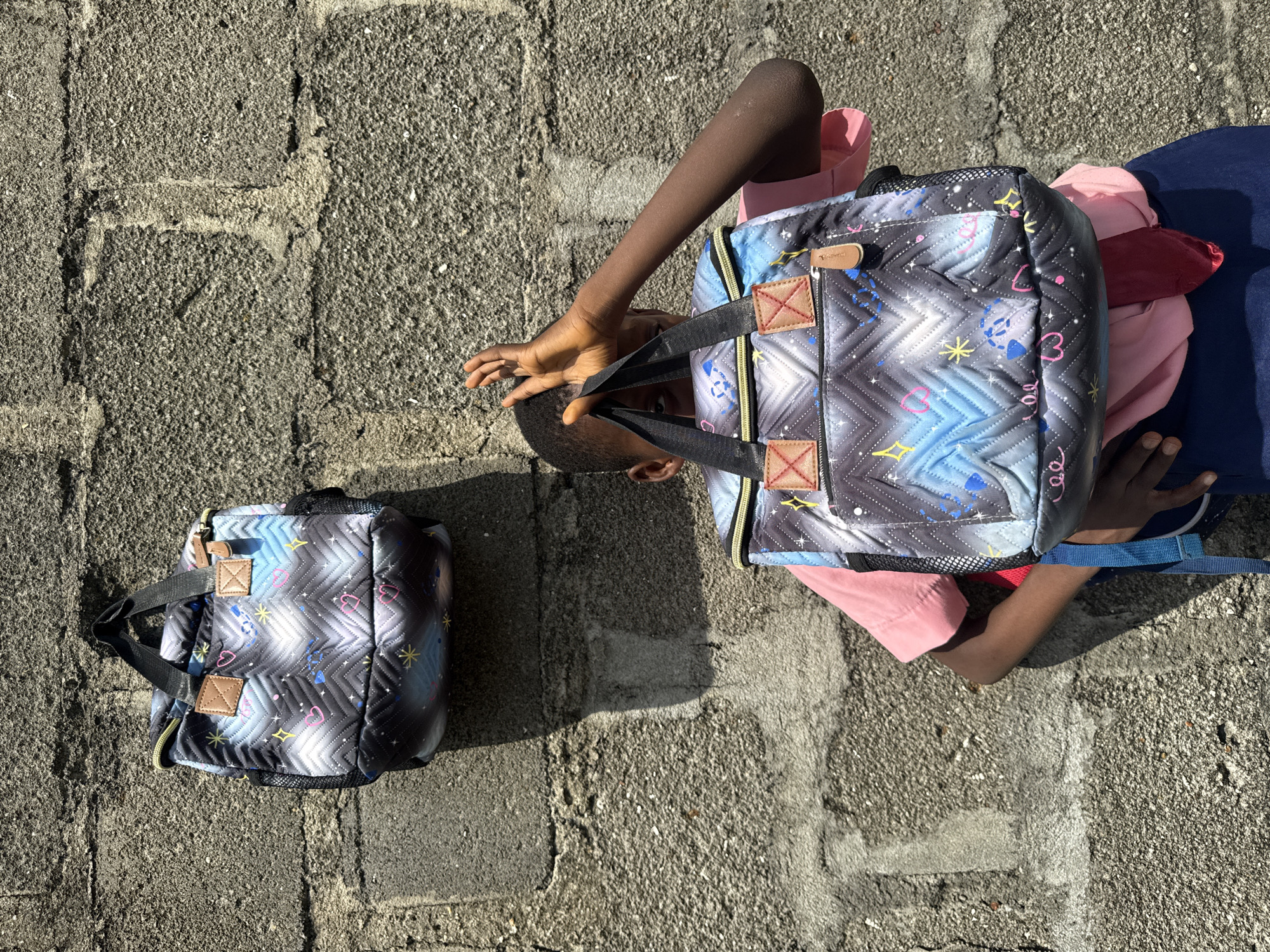 An outdoor, medium shot shows two quilted backpacks with a star and zig-zag pattern. The backpack in the foreground is held by a young Black person, who is wearing a pink t-shirt and a blue skirt. The person is mostly obscured by the backpack and is holding their hand up to their face as if to shield their eyes from the sun. The second backpack is hanging on a rough, grey concrete wall behind the person. The scene is brightly lit, casting shadows on the wall. The image appears to have been taken in Ado, Nigeria.