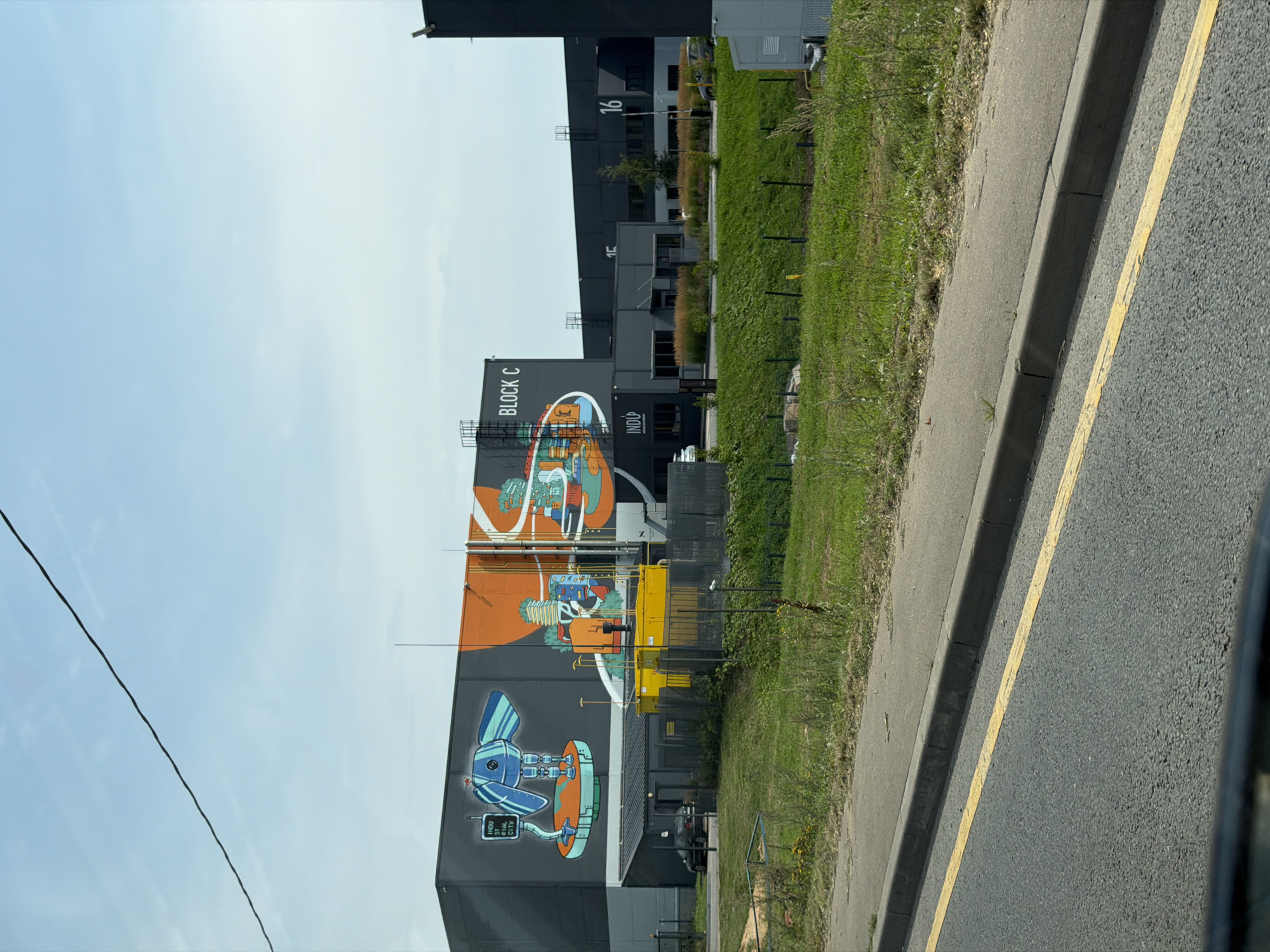 A grey industrial building is visible with a large mural on its side. The mural depicts a blue robot figure on a raised platform on the left, and a colorful abstract design with orange and green elements on the right. To the right of the mural, the text "BLOCK C" is visible. Below the mural, a smaller, dark-colored building with "INDU UP" written on it is present. Adjacent to this, a yellow structure and a black fence are visible. Further to the right, other sections of the industrial complex are apparent, with numbers "15" and "16" visible on some buildings. The buildings are situated behind a grassy slope and a paved road with a yellow line. The sky is a clear blue with a single black cable running across the top. The scene appears to be in an industrial area, possibly on the outskirts of a city, based on the presence of industrial buildings and the surrounding landscape.