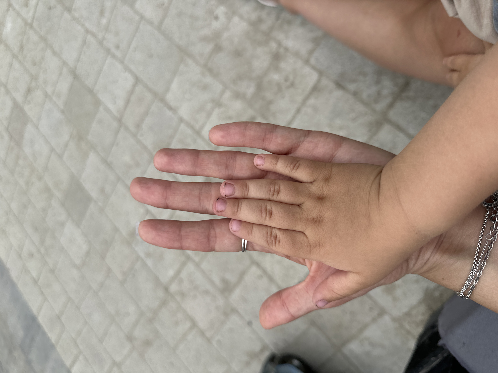 In Malinovka, Russia, two hands are superimposed against a light-colored, tiled ground. The larger hand, presumably an adult's, has a silver ring on its ring finger and displays faint dirt smudges on its palm and thumb. Placed atop this hand is a smaller, child's hand, its palm and fingers facing upward. The child's hand is positioned across the adult's palm and lower fingers, with its thumb extending slightly beyond the adult's index finger. The child's fingernails appear somewhat dirty. The scene suggests an interaction between an adult and a child, possibly a parent and child, sharing a moment of connection.