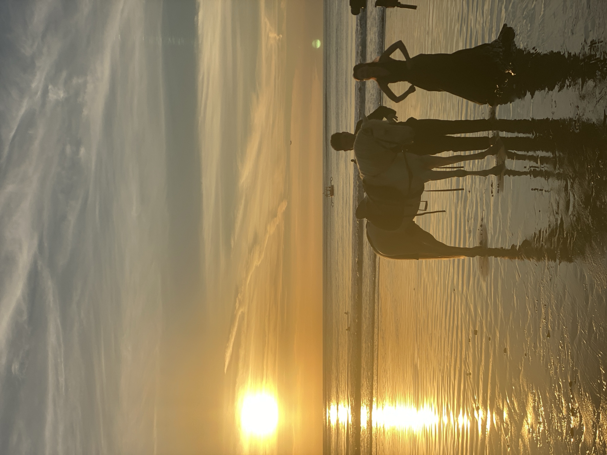 The image shows a beach scene during sunset in Kuta Selatan, Indonesia. Silhouetted figures of a woman in a dress and two individuals in casual clothing are visible, along with a white horse. The scene is set against the backdrop of a golden sunset reflected in the water. The beach appears wet, enhancing the reflective quality of the scene.