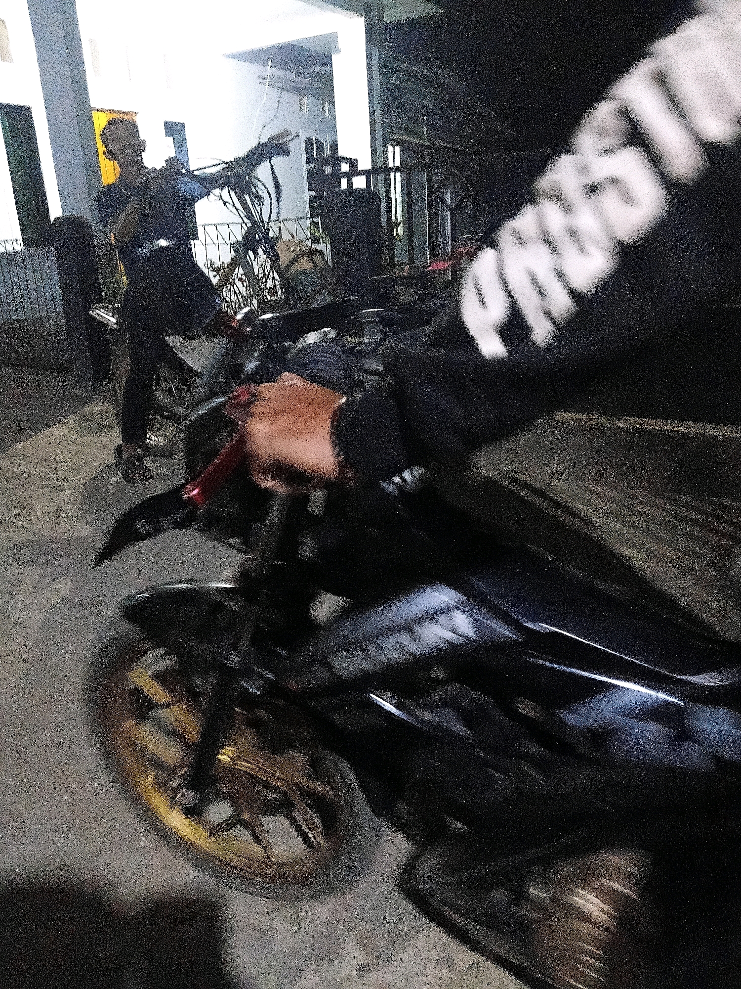 The image depicts a person in the foreground operating a motorcycle. The person is wearing dark clothing, and their hand is visible on the motorcycle's handlebar, indicating they are likely riding or about to ride the vehicle. The motorcycle itself is dark colored and has a gold-colored wheel rim. In the background, another person is visible standing near a building entrance. This area appears to be an outdoor setting, possibly a street or driveway in a residential or commercial area. The overall lighting suggests it is nighttime or dusk. Based on the location information, this scene is in the city of Sinduaji, Indonesia.