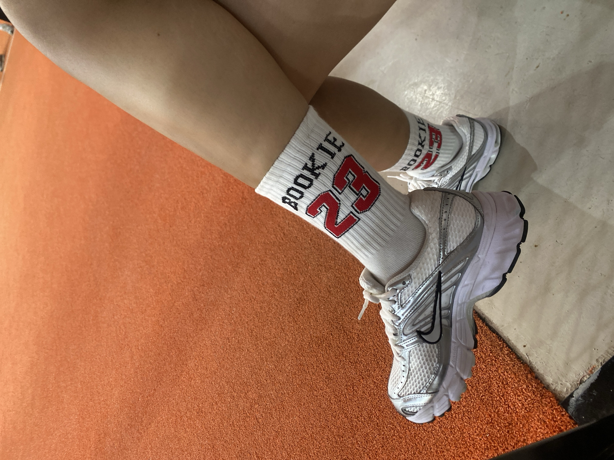 The image shows a person wearing white socks with red lettering that says "BOOKIE 23" and white and silver Nike athletic shoes. The person's legs are crossed. They are sitting on a surface that appears to be an orange, textured athletic court, possibly a tennis or basketball court. The lighting suggests an indoor environment. The image does not provide any information that could place the scene in Batam, Indonesia.