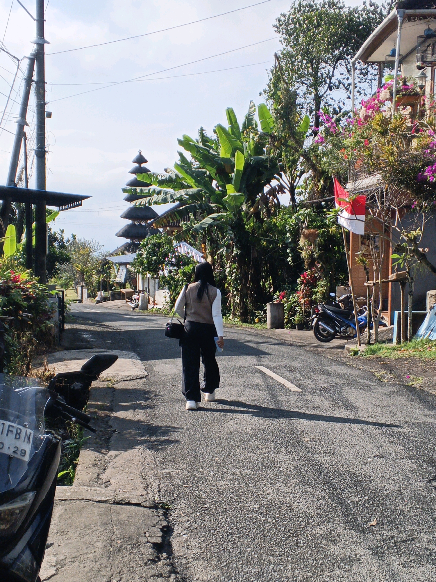 The image depicts a street scene in Penebel, Indonesia, characterized by a mixture of residential architecture and lush tropical vegetation. A woman, viewed from behind, walks away from the camera down a paved road. She is wearing dark trousers, white sneakers, and a light-colored long-sleeved shirt under a brown vest. She carries a black bag over her shoulder.
To the left of the road, a motorcycle is parked on a concrete edge, with part of its front visible. Further down the road, a distinct tiered, pagoda-like structure, likely a temple or part of a temple complex, rises above the surrounding trees. The architecture suggests a Balinese style.
On the right side of the road, a building with an orange-tiled roof is visible, adorned with blooming pink and red flowers. A red and white Indonesian flag is prominently displayed on a pole in front of this building. Another motorcycle is parked in front of the building, and what appears to be a birdcage is hanging from the eaves.
The environment is green and vibrant, with large banana leaves and other tropical plants creating a dense canopy in places. The sky is blue with some scattered clouds, indicating fair weather. The road surface is asphalt with visible tire marks and a white painted line. The overall impression is that of a warm, tropical day in a rural or semi-rural Indonesian setting.