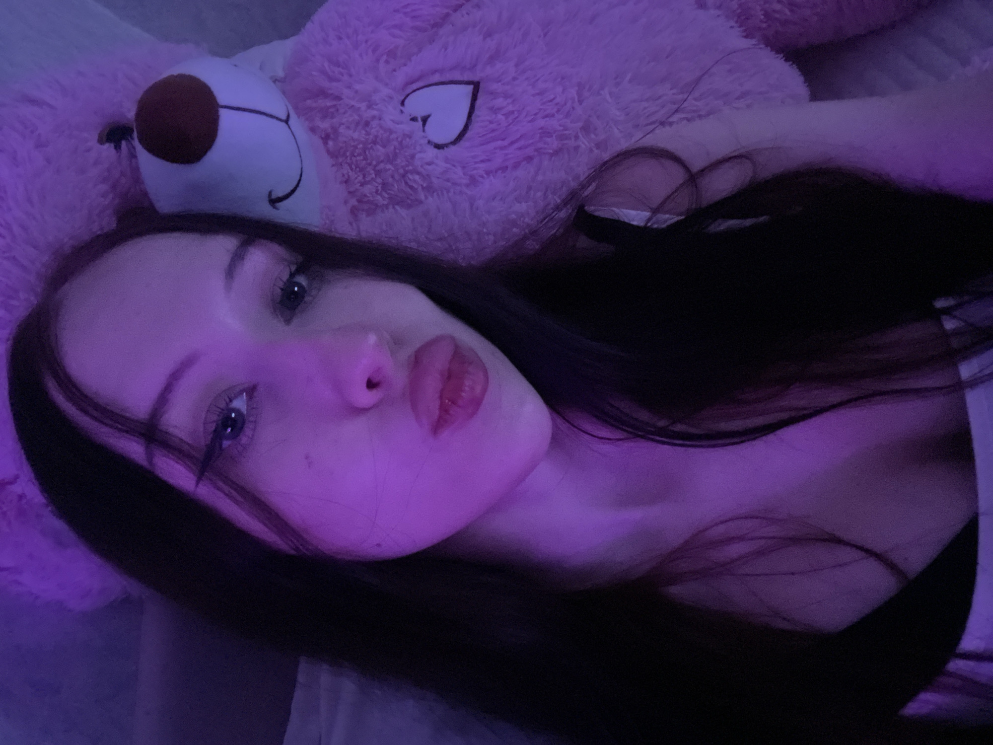 The image captures a selfie of a young adult female in a low-light, purple and pink ambient lighting environment. She has long, dark hair that falls over her shoulders. Her gaze is directed at the camera, featuring prominent winged eyeliner and long dark eyelashes framing light-colored eyes. Her lips are pursed in a "kissing" pose, appearing glossy and a reddish-pink hue. She is wearing a dark-colored top, with a white strap visible on her right shoulder/chest area. To her right, a large, fluffy pink teddy bear occupies a significant portion of the frame. The bear's head is visible, featuring a white muzzle, a brown nose, and a black stitched smile. A white outline of a heart is visible on the pink fur of the bear's chest/shoulder. The subjects appear to be resting on a soft surface, likely a bed, with some white fabric visible behind the female's head on the left side of the frame. No specific city or country can be identified from the visual information provided.