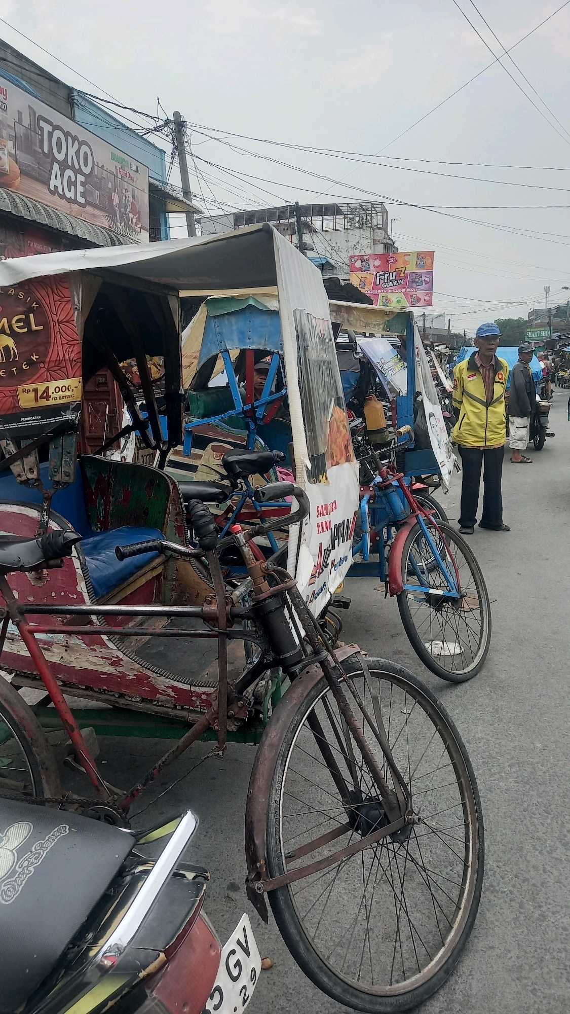 In the city of Medan, Indonesia, a street scene unfolds under an overcast sky. In the foreground, a weathered red and white pedicab, a common mode of transport in the region, stands parked. Its canopy is made of white fabric, and the seating area is covered in worn blue and green upholstery. A bicycle is attached to the front of the pedicab, its wheels and handlebars showing signs of rust and age. To the right of the pedicab, a man wearing a yellow jacket and a blue cap stands facing away from the camera. He appears to be a vendor or a driver. Behind him, more stalls and parked vehicles, including motorbikes, line the street. A large billboard advertising "TOKO AGE" is visible in the background, along with various other signs in Indonesian. The road surface is paved, and power lines crisscross the sky. The overall impression is one of a busy, bustling street market or transportation hub in a Southeast Asian city.
