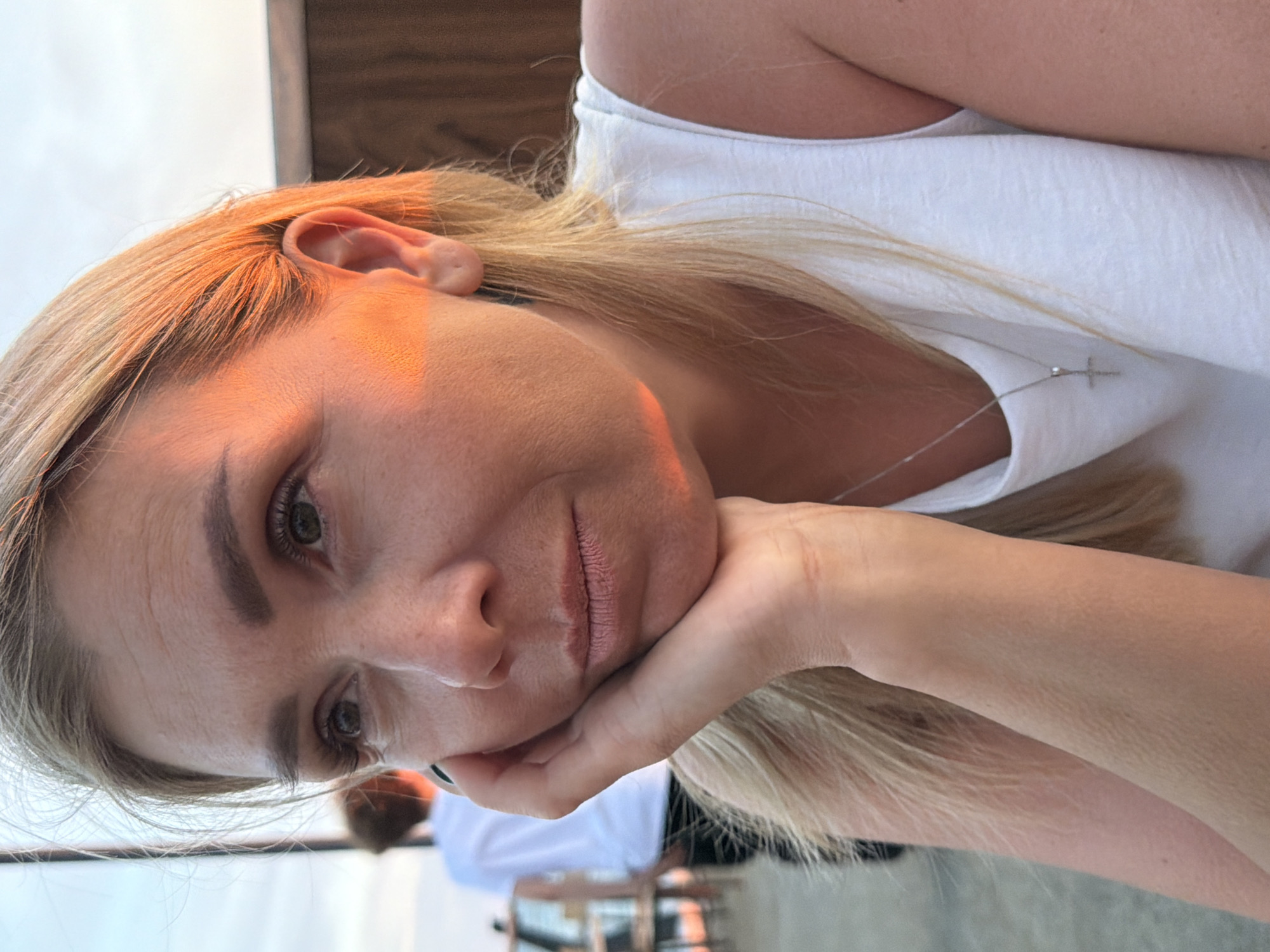 The image is a close-up portrait of a Caucasian woman with blonde hair, who is resting her chin on her hand. She is wearing a white tank top and a silver cross necklace. Her eyes are looking to the left, and the setting sun casts an orange glow on her face and hair. In the background, a blurred figure in a white shirt can be seen. The setting appears to be outdoors, possibly a restaurant or public area, with some wooden structures visible. The woman's facial expression is pensive.