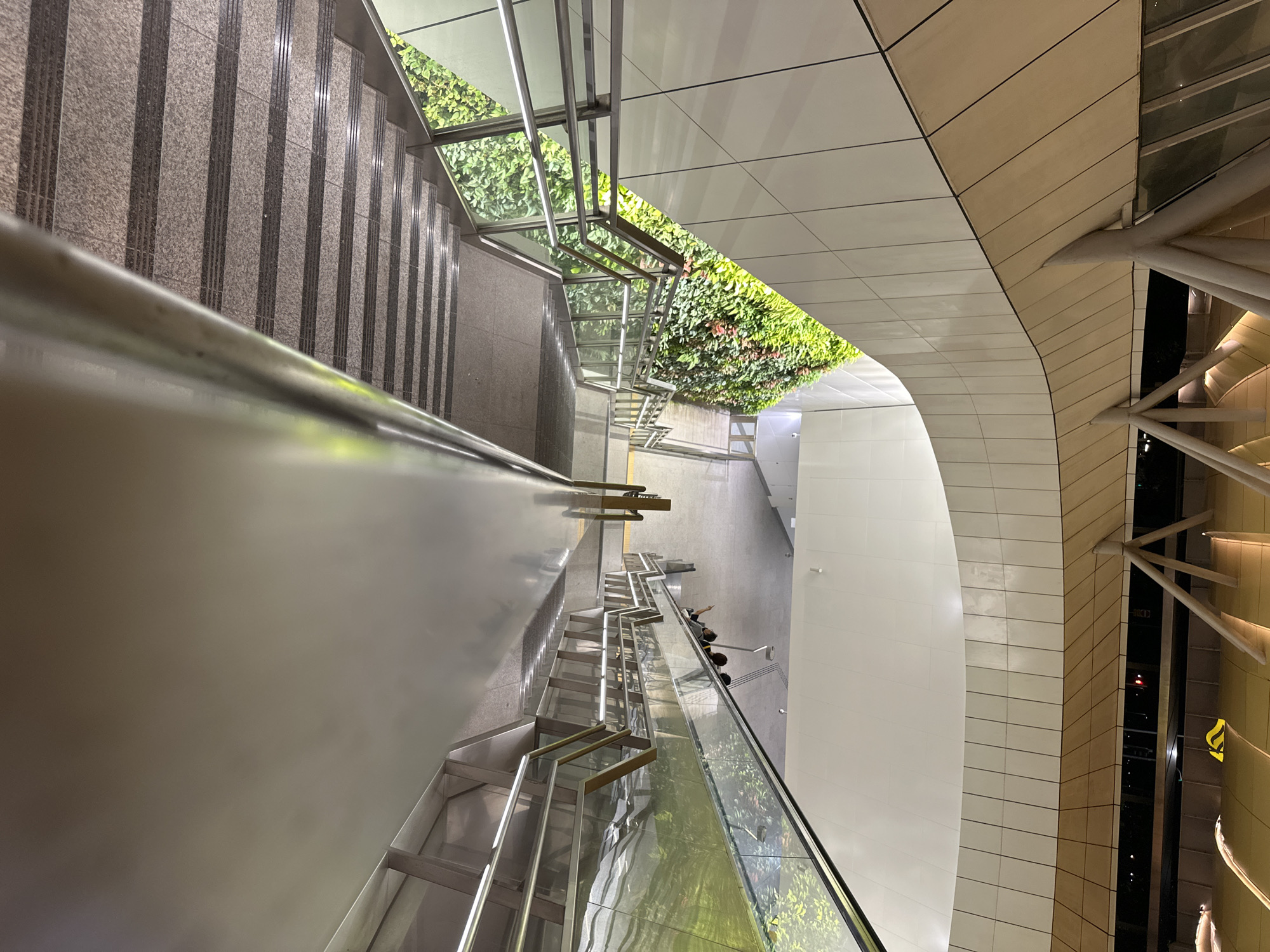 The image captures a downward view of an escalator and a staircase in a modern urban setting. A vertical garden adorns the wall to the left of the stairs, featuring lush green foliage with hints of red flowers. The escalator to the right showcases a reflective surface, mirroring the greenery and the surrounding architecture. Several individuals are visible on the escalator, some standing and others in conversation. The staircase is constructed with granite steps and metallic railings, illuminated by embedded lights. The ceiling above features a distinctive architectural design with curved panels and supporting beams, suggesting a covered walkway or transit hub. The overall environment is clean and contemporary, indicative of public infrastructure in a well-maintained city. The scene likely depicts an area within Singapore, given the prevalence of such modern green architecture in public spaces and transit facilities in the city-state.