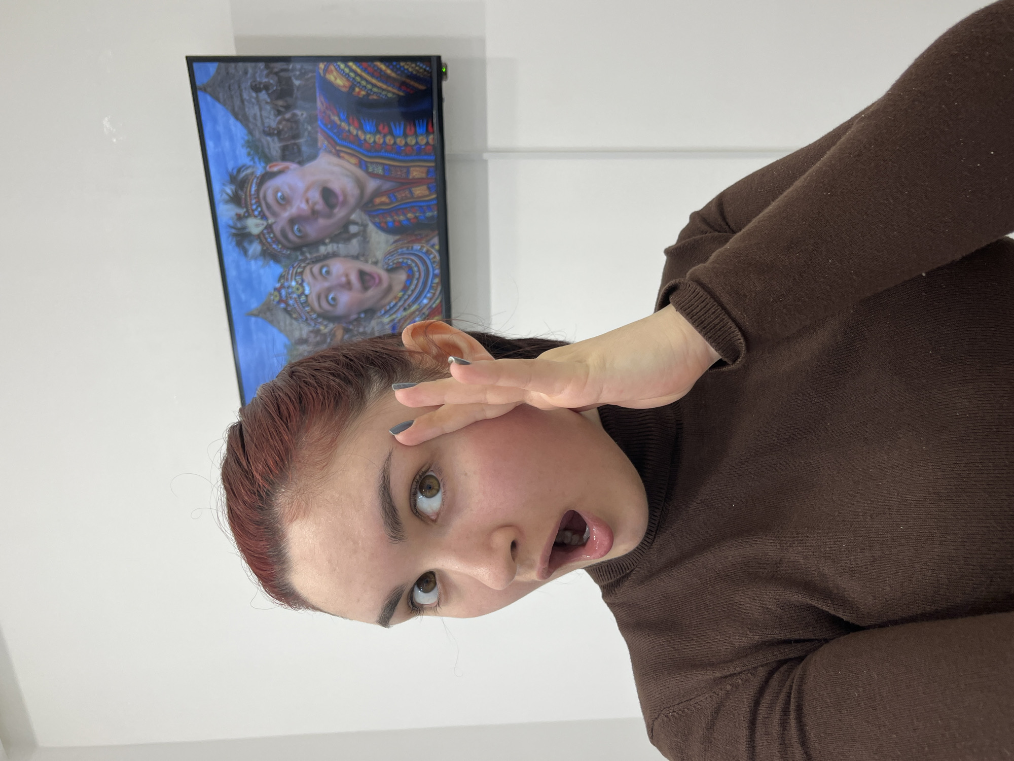 The image captures a female subject indoors in Neftekamsk, Russia, presented in a close-up perspective. She possesses fair skin, dark reddish-brown hair pulled back, and light brown eyes. Her facial expression conveys surprise, characterized by wide-open eyes and an agape mouth. Her left hand is positioned near her left ear, fingers slightly spread, with dark grey nail polish visible on her fingernails. She is attired in a brown long-sleeved turtleneck sweater.
Mounted on a plain white wall behind the subject, to her right, is a black-framed flat-screen television. The screen displays an image or video featuring a male and a female individual, both exhibiting exaggerated expressions of surprise, mirroring the primary subject's demeanor. These individuals on screen are adorned in colorful, patterned clothing and elaborate headwear, suggestive of tribal or ceremonial attire. The background on the television includes hut-like structures and indistinct figures. A green indicator light is visible on the bottom right bezel of the television, and a white cable extends vertically downwards from behind the unit. The surrounding wall surface is uniformly white.