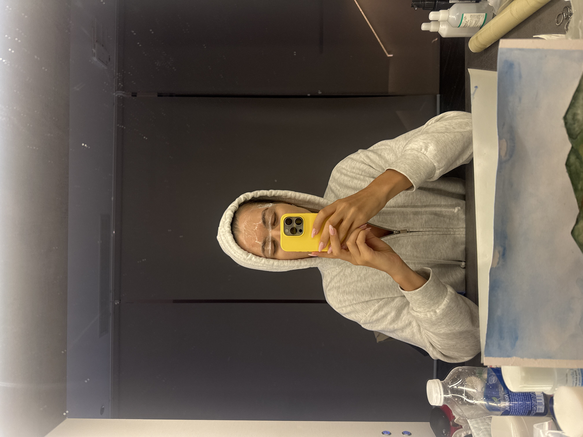 A person is standing in front of a mirror, wearing a gray hooded sweatshirt. The person is holding a yellow smartphone in their hands and appears to be taking a selfie. There is a white cream-like substance applied to the person's face, particularly around their eyebrows. The background includes a dark wall or surface, possibly part of a dressing room or studio setup. Various objects like bottles, tubes, and papers are present on a surface or shelf near the mirror.