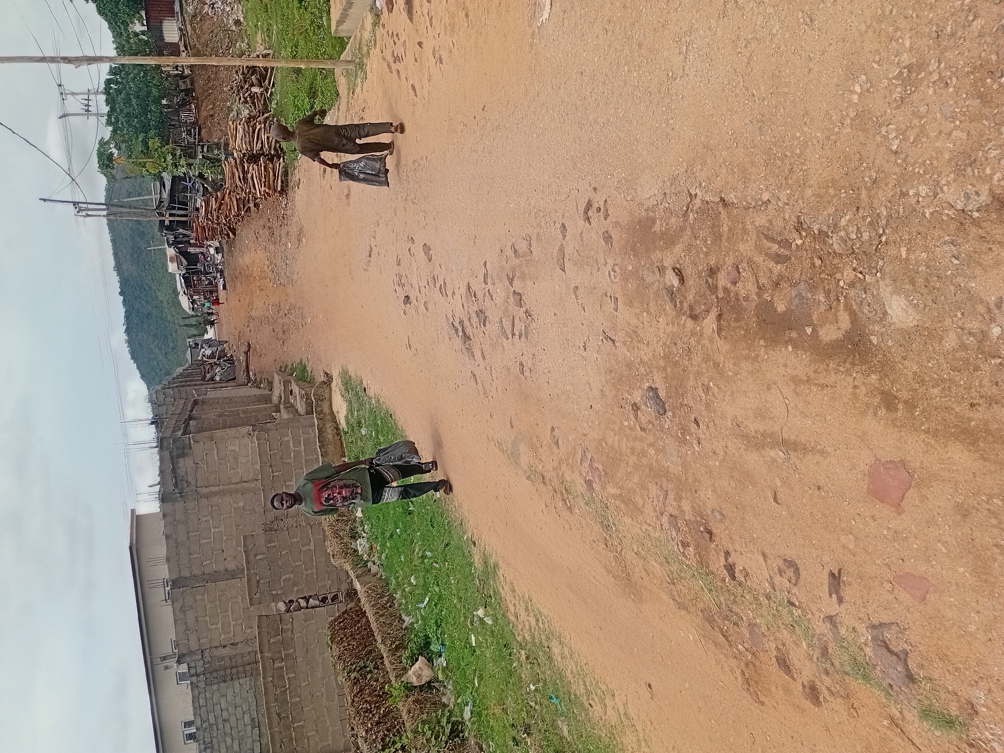 The image shows a dirt road in Unknown, Unknown. On the left side of the road, there is a building under construction. On the right side, there is vegetation. Two people are walking on the road. One is closer to the building and the other is further up the road. A mountain is visible in the background. Electrical poles are visible on the left side of the image.