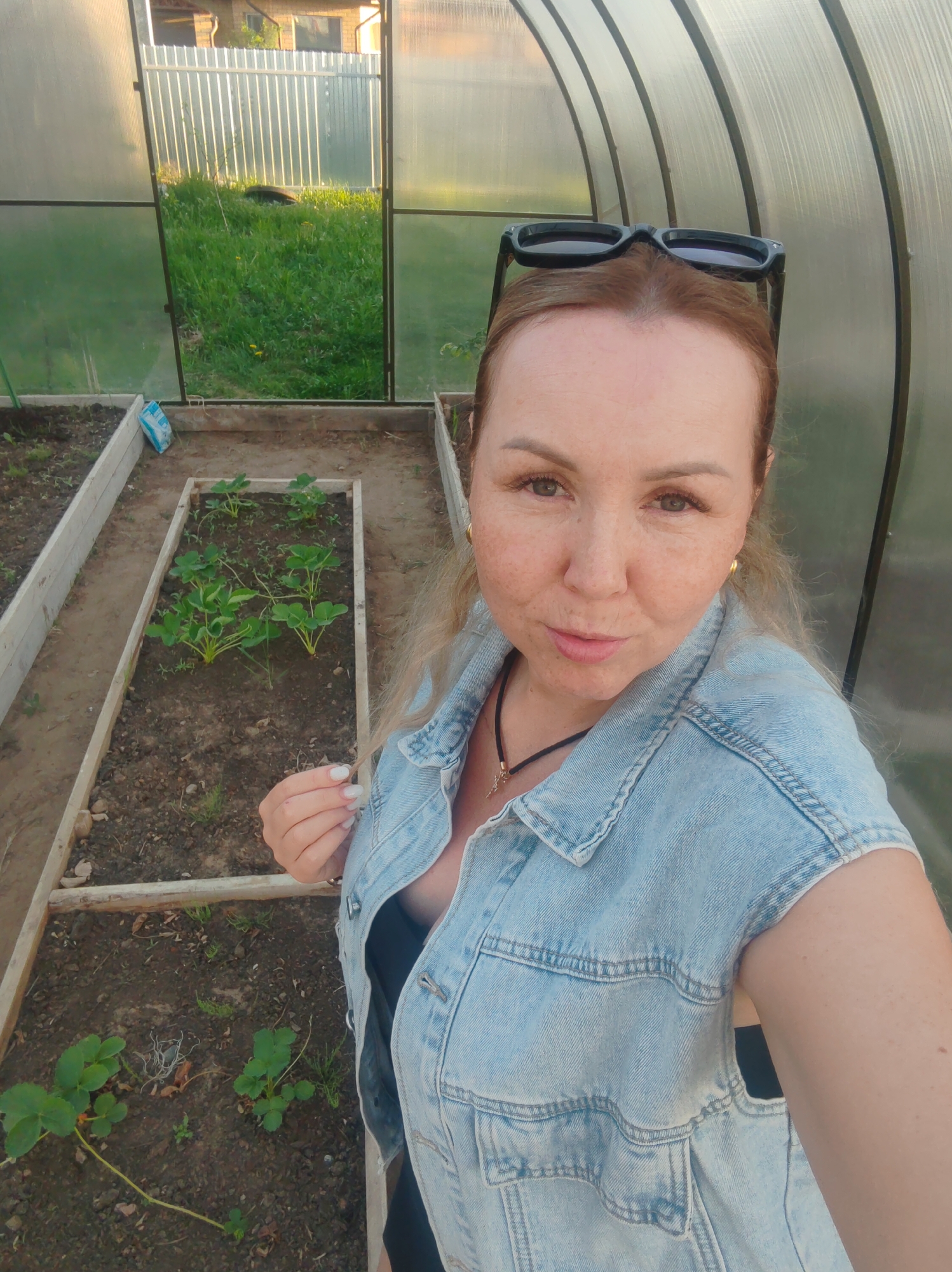A selfie shows a woman in a greenhouse in Chaikovsky, Russia. She wears a sleeveless denim shirt over a black top and has sunglasses on her head. Inside the greenhouse, there are raised garden beds with various plants, including strawberries. Outside the greenhouse, grass and a fence are visible.
