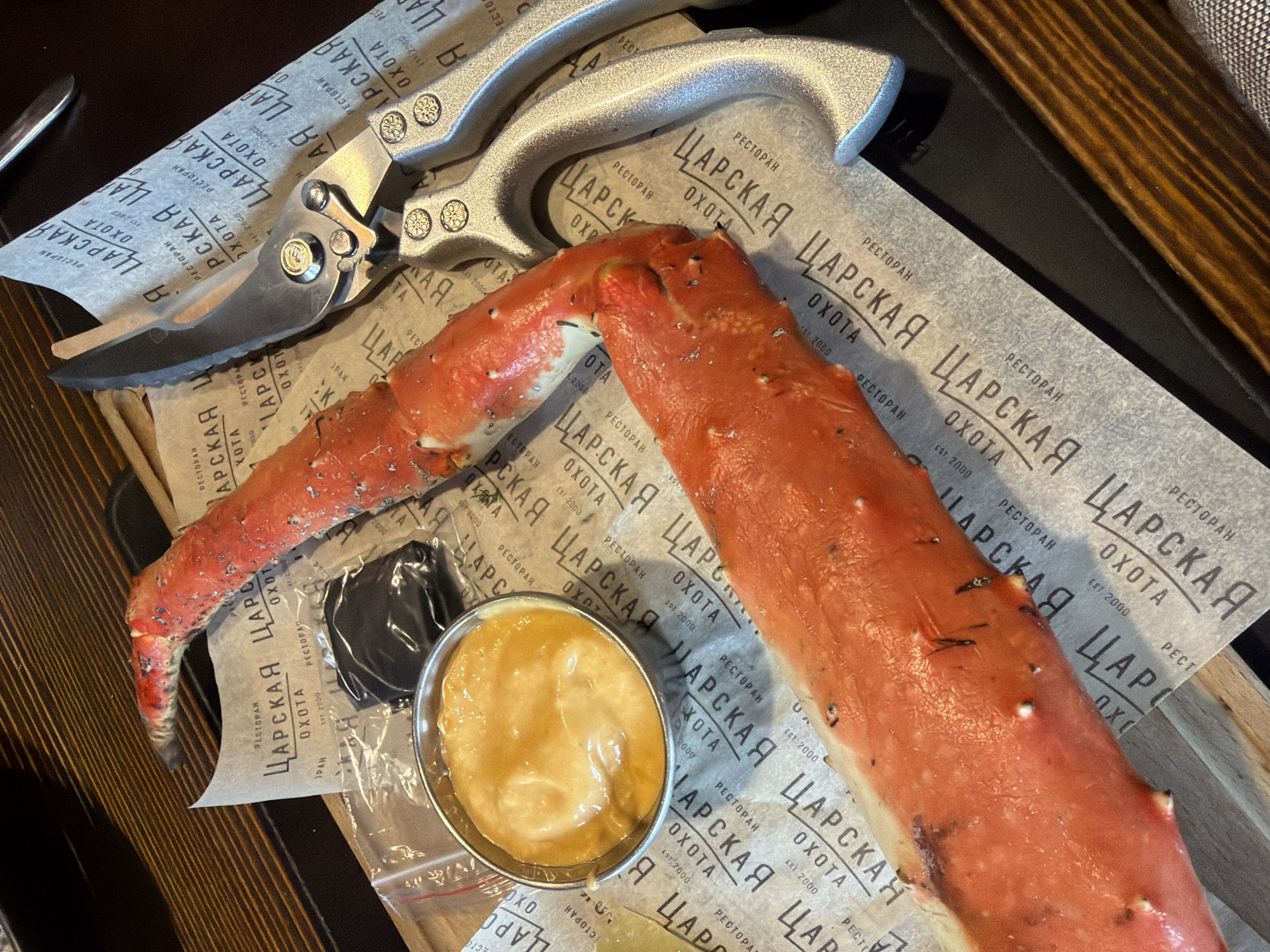 The image displays a large crab leg served on a dark wooden surface, possibly a table. The crab leg is bright red and rests on a piece of patterned paper. Next to the crab leg is a small metal ramekin filled with a yellowish sauce. A pair of silver kitchen shears with serrated edges is positioned on top of the patterned paper, near the crab leg and ramekin. A small plastic bag containing a dark substance, possibly a condiment or seasoning, is also visible. The background shows the edge of a dark tray and a wooden table with a visible grain. The overall impression is that of a meal being prepared for consumption. The text on the paper appears to be in Cyrillic. Without additional information or context, it is not possible to determine the specific location in Murmansk, Russia.