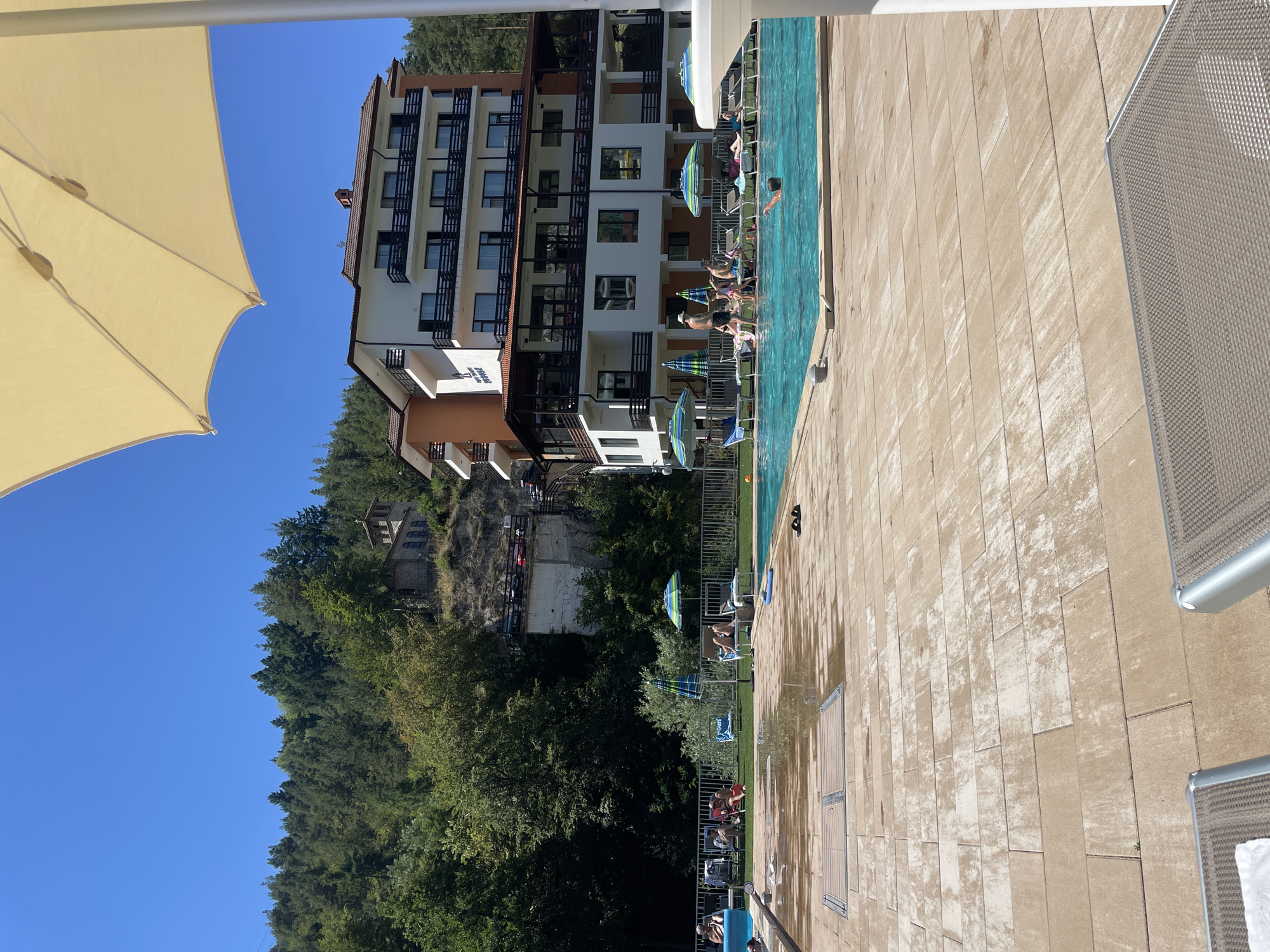 This is a wide shot of an outdoor swimming pool area at a resort in Banite, Bulgaria. In the background, a multi-story hotel building with balconies and windows is situated against a backdrop of a dense, green forest and a steep, rocky incline with a small stone structure on top. The hotel appears to be a light color with orange accents. In the foreground, the pool is filled with bright blue water, and several people are seen lounging on chairs or swimming. The pool deck is paved with large, tan rectangular tiles, some of which are wet, indicating recent use or cleaning. Several beach umbrellas are set up around the pool area to provide shade. The sky is a clear, vibrant blue.