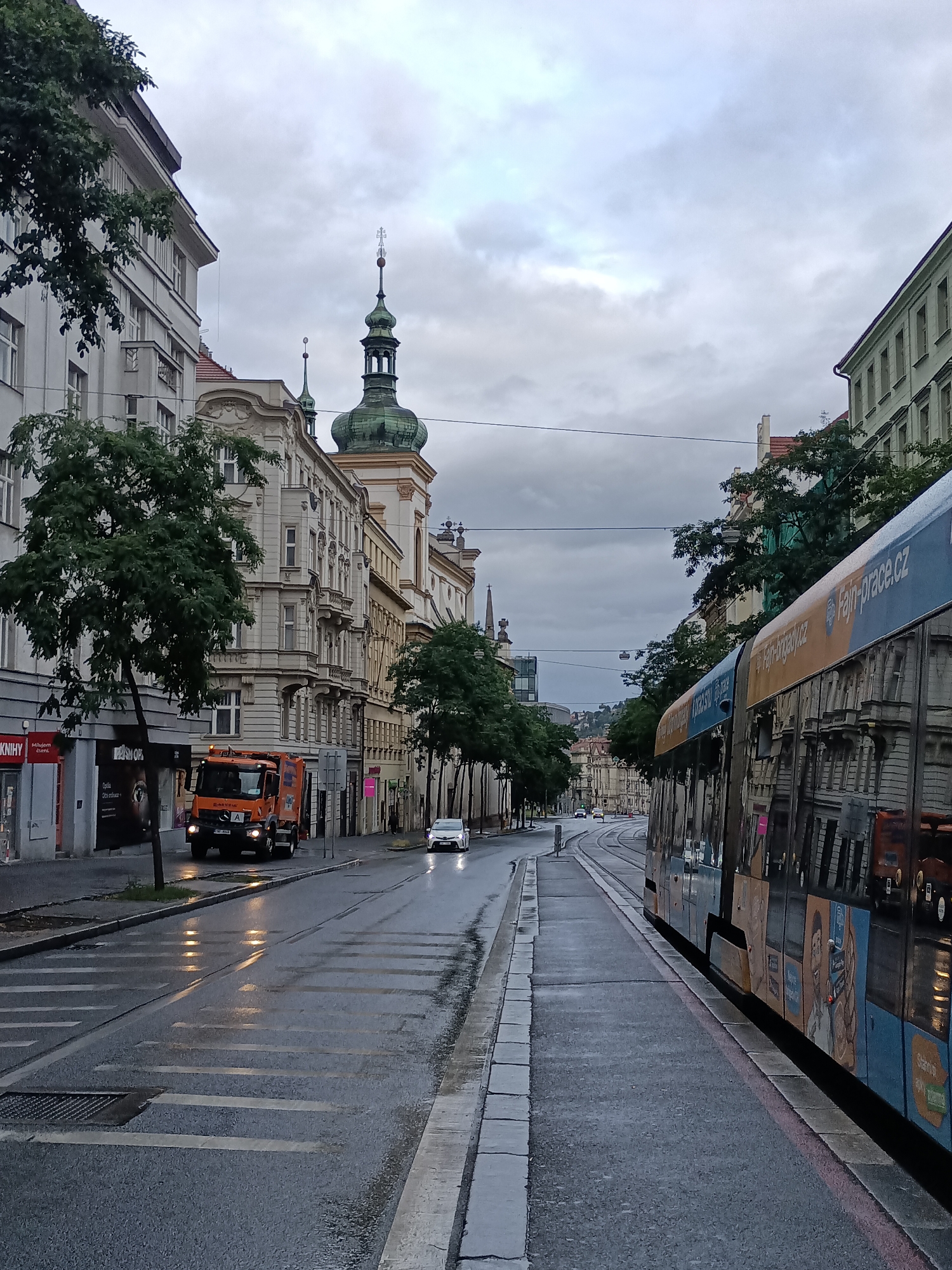 The image depicts a street scene in Prague, Czechia, on a cloudy day, possibly after rain, as evidenced by the wet asphalt and reflections. A tram with colorful advertisements runs along the right side of the street, its windows reflecting the surrounding buildings. On the left side, an orange refuse collection truck is stopped near the curb. Behind the truck, pedestrian crossings are visible on the wet road. The street is lined with multi-story buildings featuring architectural details typical of European cities, including balconies and ornate facades. A prominent green-domed tower with a cross on top rises above the buildings in the background. Trees line the sidewalks, some with branches extending over the road. The sky is overcast with dramatic cloud formations. The overall impression is that of an urban environment, likely a city center or a busy thoroughfare, captured during a period of subdued natural light.