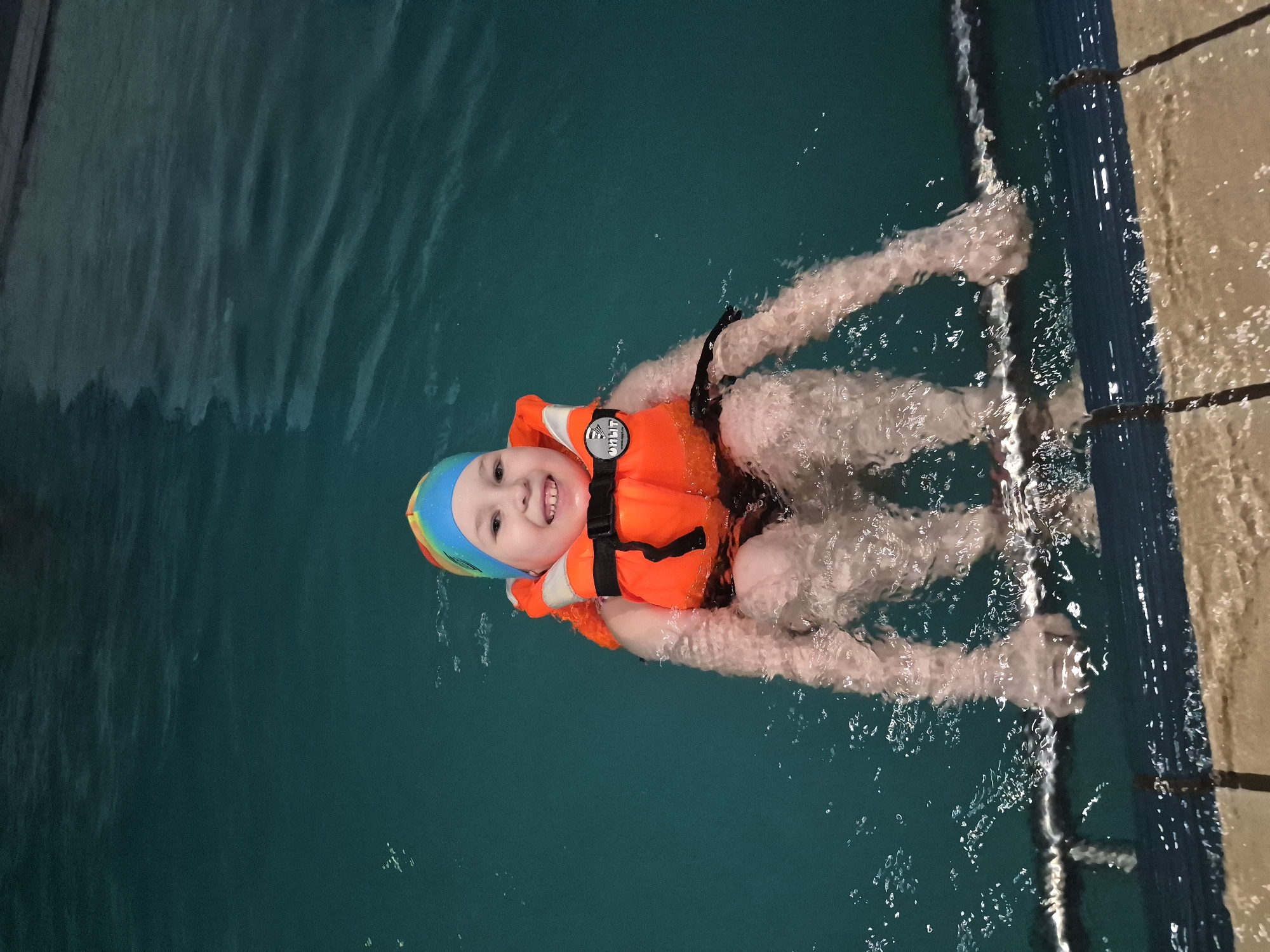 A young child wearing an orange life vest and a multi-colored swim cap is floating in a swimming pool. The child is smiling and looking at the camera. Their hands are gripping the side of the pool. The water is clear and green. The pool is in Tobolsk, Russia.