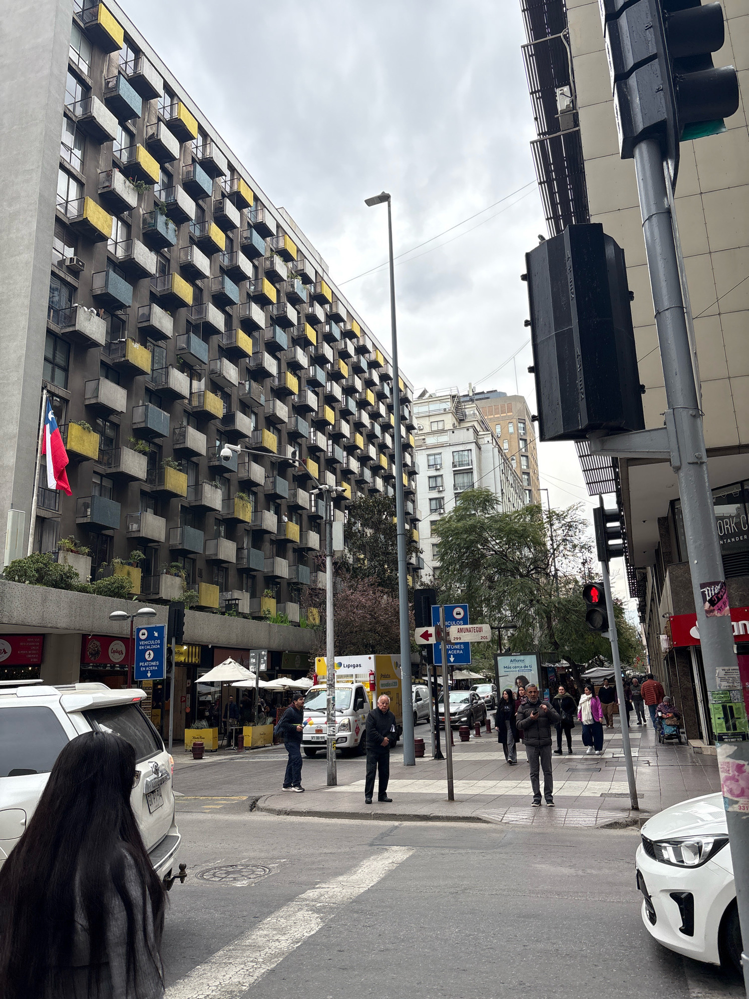 The image captures an urban street scene in Santiago, Chile, likely on a cloudy day. In the foreground, a white SUV is parked facing away from the viewer, and the back of a person with long dark hair is visible, facing the street. To the left, a multi-story building with a distinctive facade of numerous balconies, alternating in yellow and blue colors, dominates the view. A Chilean flag is mounted on the left side of this building.
The street features crosswalks and traffic signals. A traffic light is prominently displayed on the right side of the image, showing a red light. Several people are seen walking on the sidewalk and crossing the street. Some are dressed in jackets and casual wear, suggesting a temperate climate. Businesses with signs like "Lipigas" and "Afíllatex" are visible along the street. The architecture suggests a mix of older and newer buildings. The overall impression is of a busy urban intersection in a South American city.