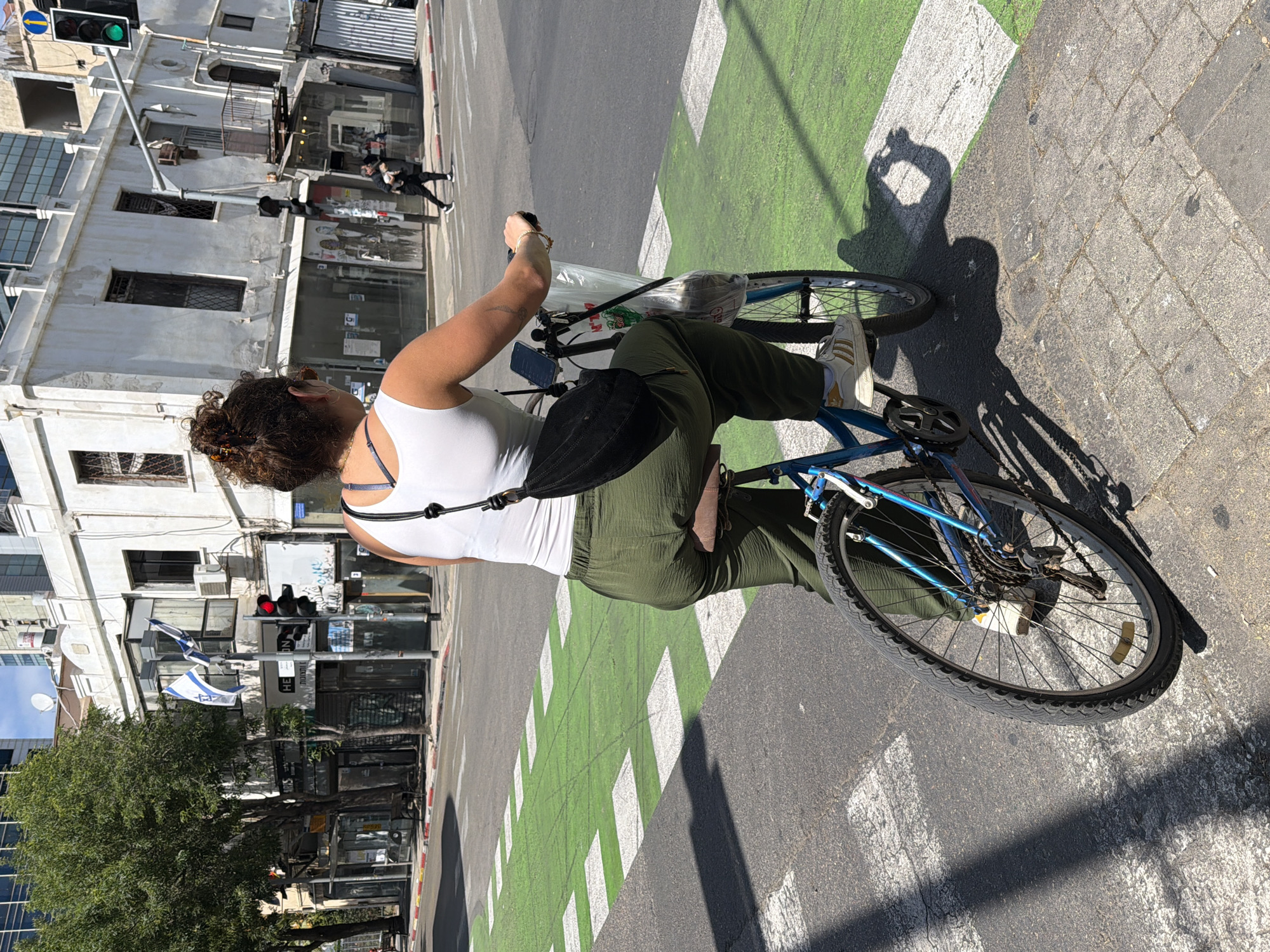 A woman is riding a blue bicycle on a street with green and white crosswalk markings. She is wearing a white tank top and green pants. A black bag hangs across her body. The traffic light is green. People are walking on the sidewalk in the background. Israeli flags are displayed in front of buildings. The location is Tel-Aviv, Israel.
