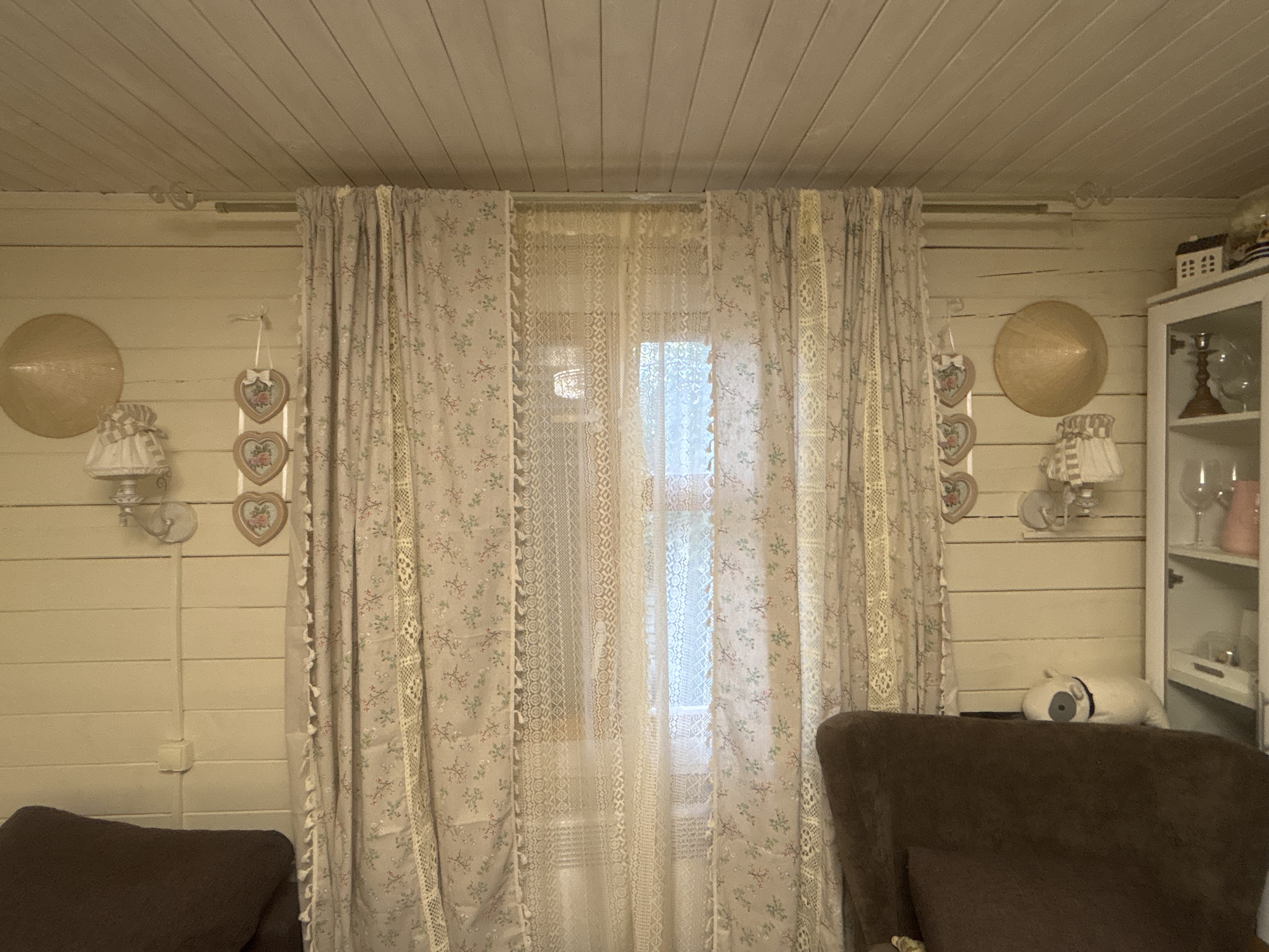 The image depicts an indoor scene, likely a living room, in a house located in Лотос Снт (марьино), Russia. The room has a rustic aesthetic, with light-colored wooden paneling on the walls and ceiling. In the center, a window is covered by floral patterned curtains and a white lace curtain. To the left of the window, there are two decorative heart-shaped ornaments hanging on the wall, a conical straw hat, and a wall-mounted lamp. To the right of the window, there is a white cabinet with glass shelves displaying decorative items, including glassware and a small white dog figurine. A dark brown armchair is partially visible in the foreground on the left and right sides of the image. The overall impression is one of a cozy, perhaps somewhat dated, interior.