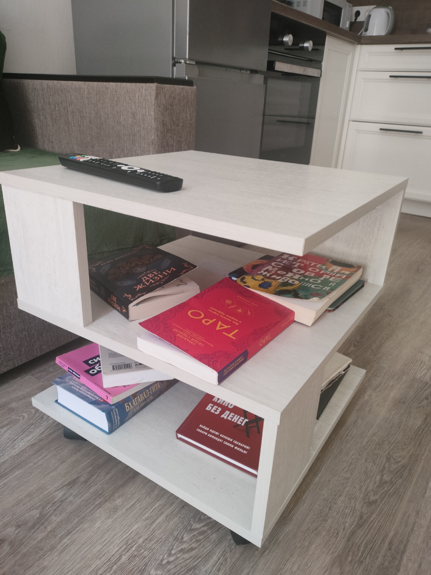 A white coffee table with multiple shelves stands on a wooden floor in an indoor setting. On the top shelf of the table is a black remote control. The shelf below it contains two books: one dark-covered book with Russian text and a lighter-colored book underneath it. The third shelf from the top holds a red book titled "ТАРО" and another book with a colorful, abstract cover and Russian text. The bottom shelf displays a pink book, a blue book with Russian text, and a red book with Russian text on its cover. The background reveals a textured couch and a glimpse of a kitchen area with white cabinets and appliances. The scene is consistent with an apartment in Lyubertsy, Russia, a suburban city within the Moscow Oblast.