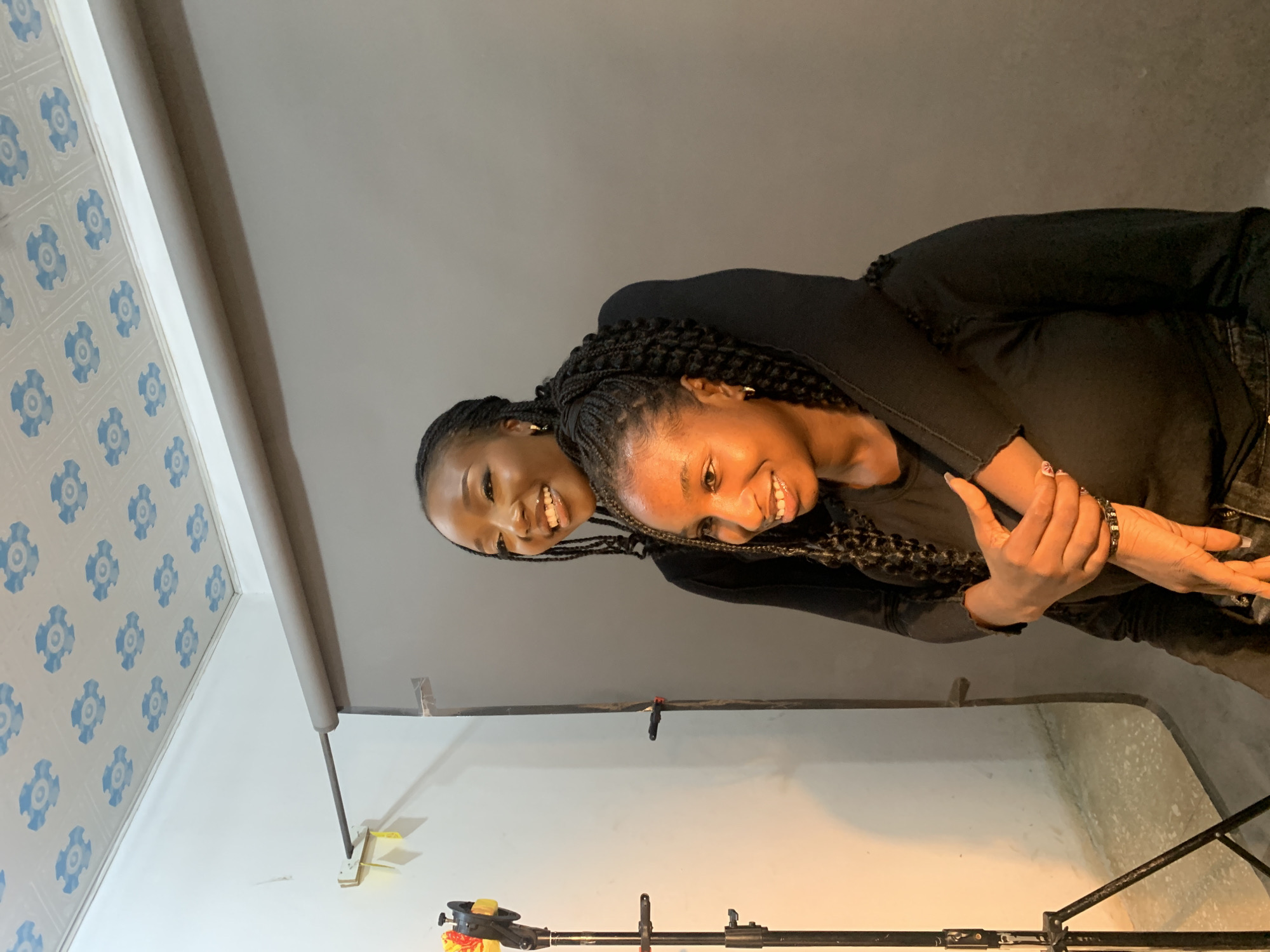 Two women with braided hair, wearing black tops, are posed in an indoor studio environment. One woman is giving the other a piggyback ride. A photography backdrop and lighting equipment are visible in the background. The location is suggested to be Gura Top, Nigeria, but no details in the image confirm that.