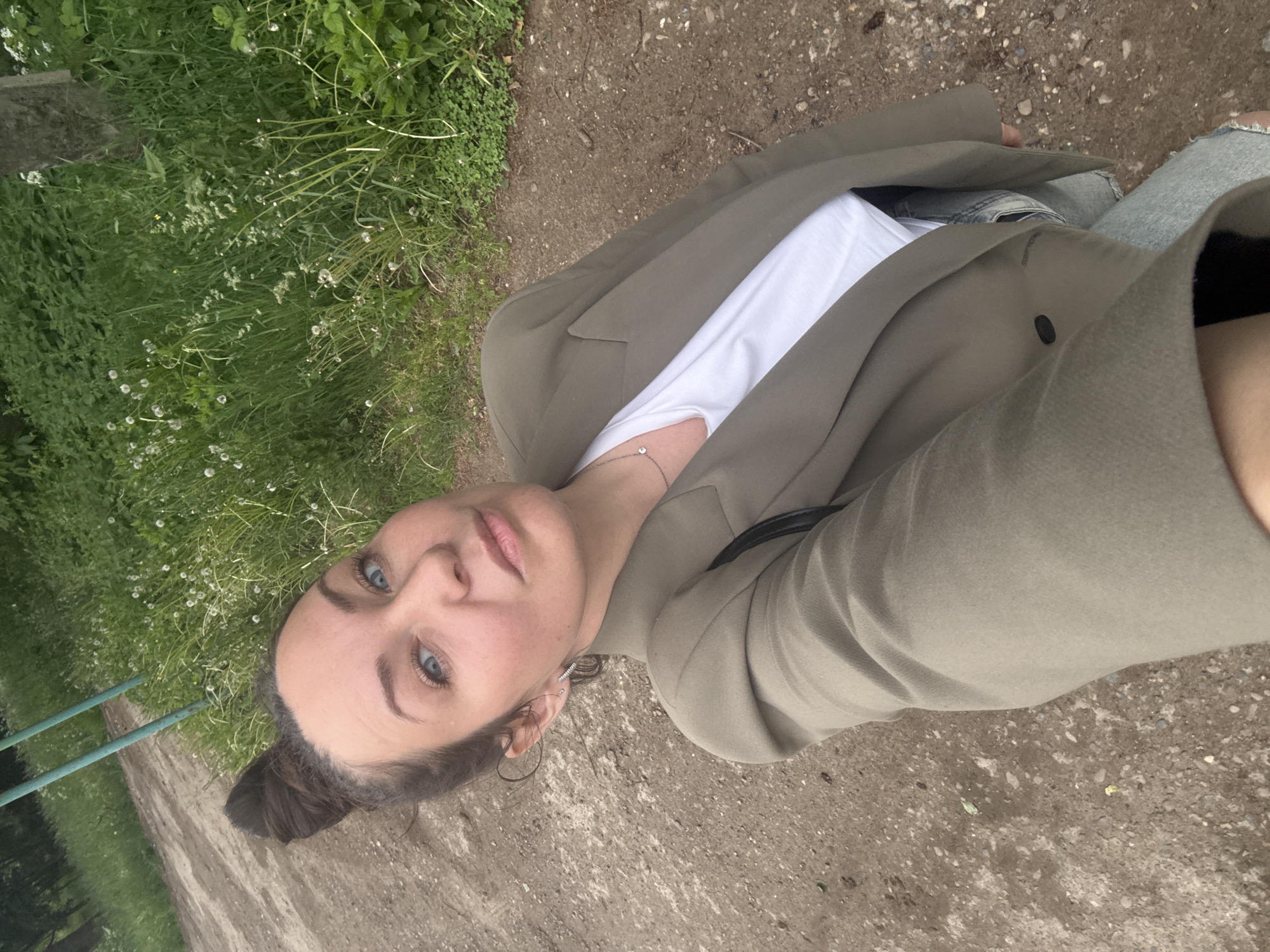 The image is a selfie of a young adult female taken outdoors in Chaikovsky, Russia, during what appears to be an overcast day. The woman has light skin, striking blue eyes, and dark hair tied back into a bun. She gazes directly into the camera with a neutral to slightly contemplative expression.
She is dressed in a casual yet put-together outfit, consisting of a white t-shirt, a sage green or olive blazer, and light-wash ripped jeans. A delicate silver necklace featuring a small, clear gemstone is visible around her neck.
The setting is a natural environment. To her right, there is a rough dirt path or road. To her left, the ground is covered in lush green vegetation, including grass and various leafy plants, some adorned with small white, spherical seed heads, possibly from dandelions. In the background on the far left, two parallel green metal bars are partially visible, suggesting a fence or gate. The lighting is soft and diffused, indicating an overcast sky without harsh shadows. There is no visible text in the image.