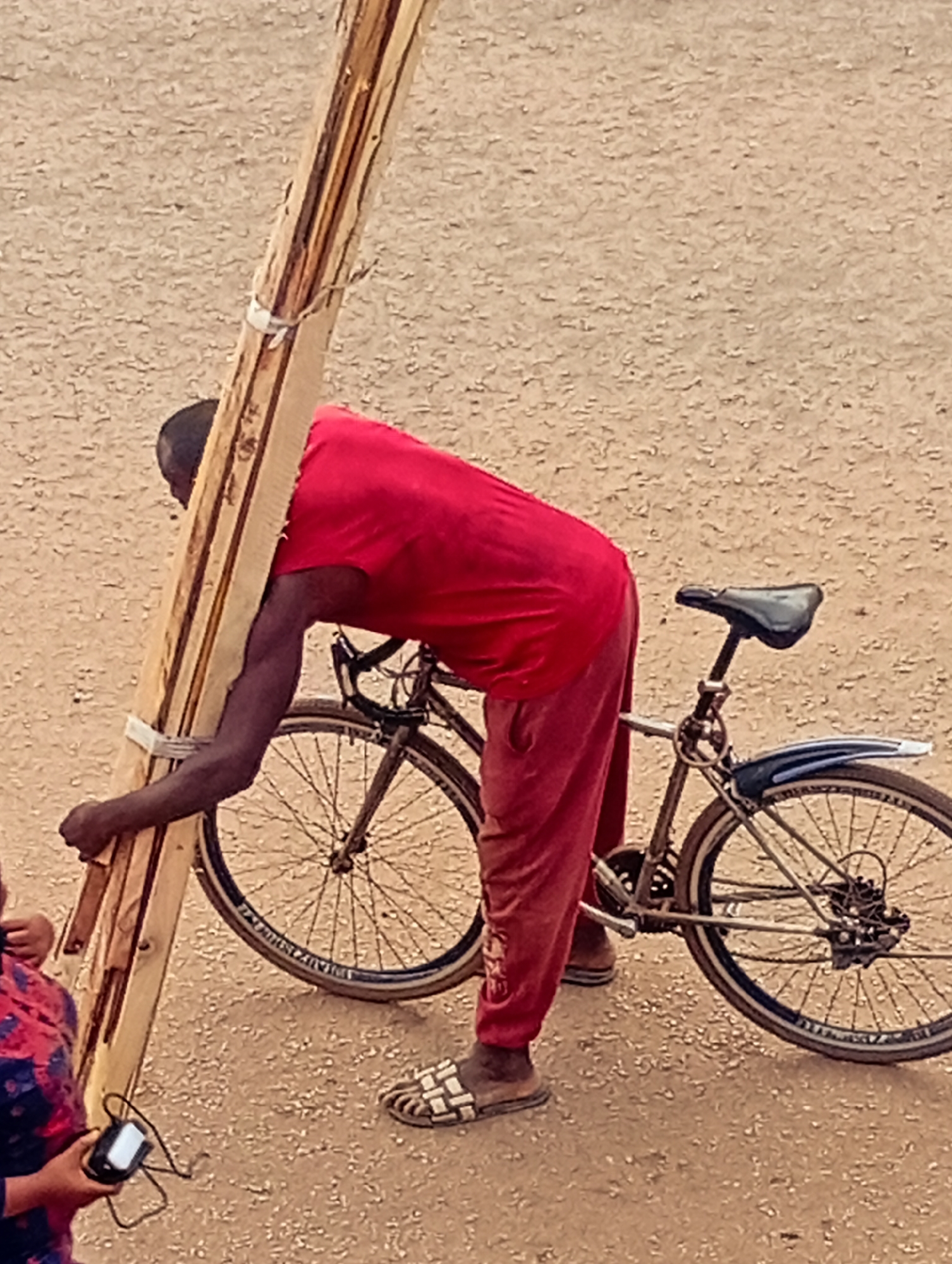 In Bukuru, Nigeria, an African man in a red shirt and pants is carrying a bundle of wooden boards on his left shoulder. The boards are tied together with white ties. He is next to a black bicycle. The man is wearing sandals. In the foreground, a person partially visible is holding a device with a screen and buttons. The ground appears to be a dirt road.