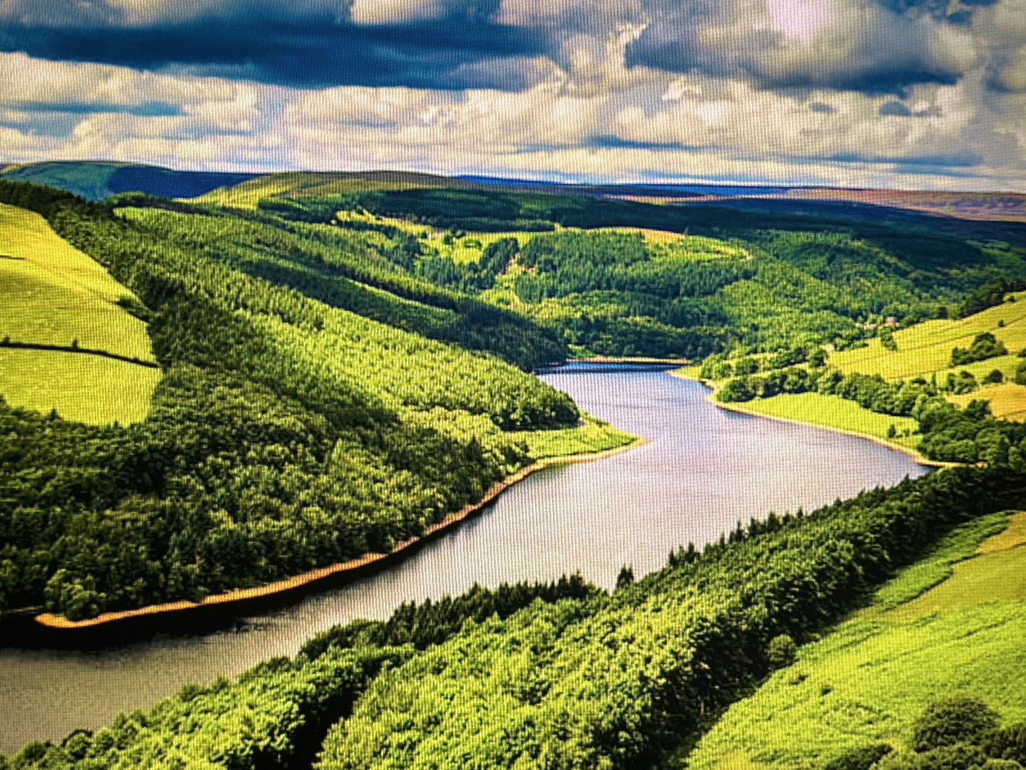 The image depicts an aerial view of a vast, verdant landscape dominated by rolling hills, a winding river, and a dramatic sky. Lush green forests cover the slopes of the hills, interspersed with patches of bright yellow fields. The river, a calm, reflective body of water, snakes through the valley, its banks lined with dense trees. In the distance, layers of hills recede into the horizon, merging with a sky filled with dark, textured clouds. The scene evokes a sense of natural beauty and tranquility. There is no indication that this location is in Sofia, Bulgaria.