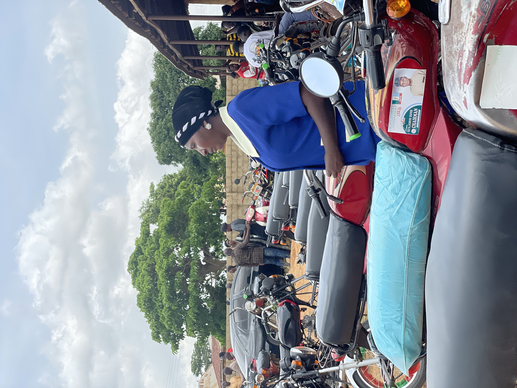 The image depicts an outdoor scene in Kassa, Nigeria, characterized by a cloudy sky and a gathering of people around numerous motorcycles. In the foreground, several motorcycles are parked, with their seats and handlebars visible. One red motorcycle prominently features a campaign poster with the text "GOODWILL 2026" and an image of a man identified as "BENJAMIN DUNG, APC CHAIRMAN". A woman wearing a blue dress and a black headwrap with white trim stands to the left, looking to the right. Behind the motorcycles, more people are gathered, some standing near a dark-colored car and a rustic wooden structure. Trees and buildings are visible in the background, suggesting a semi-urban or rural setting within Kassa. The overall atmosphere appears to be one of a community event or political gathering.