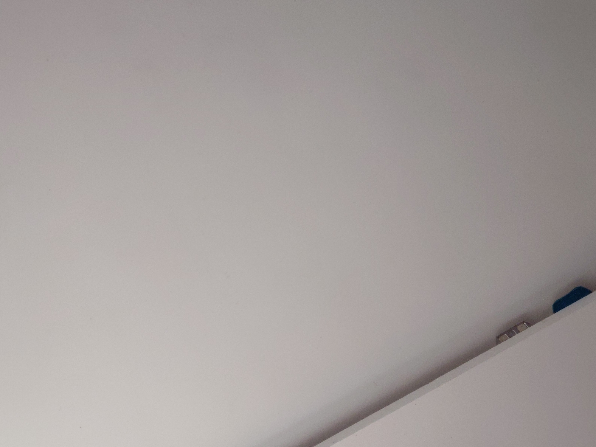 The image captures a close-up, angled view of an interior space, dominated by a large, smooth, light grey or off-white surface, likely a wall or ceiling. This surface fills the majority of the frame.
A distinct diagonal line extends from the lower-left center towards the upper-right corner, marking the edge of a different, brighter white surface in the foreground, occupying the bottom right portion of the image. This brighter surface appears to be a flat, clean edge, possibly part of a cabinet, shelf, or another piece of furniture.
Along the intersection of these two surfaces, near the right edge of the frame, several small, metallic objects are visible. These appear to be small, shiny fasteners, clips, or parts of a hinge mechanism. Immediately to their right and partially obscured, a small section of a solid, dark blue object can be seen.
No people or activities are present. The lighting is soft and uniform, suggesting indoor ambient light. There are no visible texts or specific details within the image itself that directly indicate its location in Santa Ana, Venezuela.