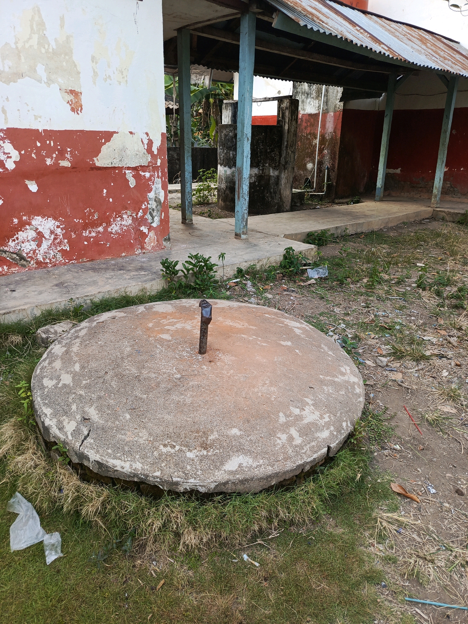 The image shows an outdoor scene in a somewhat dilapidated area. In the foreground, a large, circular concrete manhole cover with a metal handle is prominent. The cover shows signs of wear and a crack. It is surrounded by dry and green grass, with some scattered leaves and debris. Behind the manhole cover, there is a building with peeling red and white paint on its walls. A covered walkway with blue support poles and a corrugated metal roof extends from the building. Various structures, including concrete tanks and pipes, are visible in the background. The ground is a mix of dirt, grass, and scattered refuse. The overall impression is of an unkempt, possibly abandoned, or functional but neglected outdoor space. The context of Megang Sakti II, Indonesia, suggests this could be a public utility area or part of a community facility.
