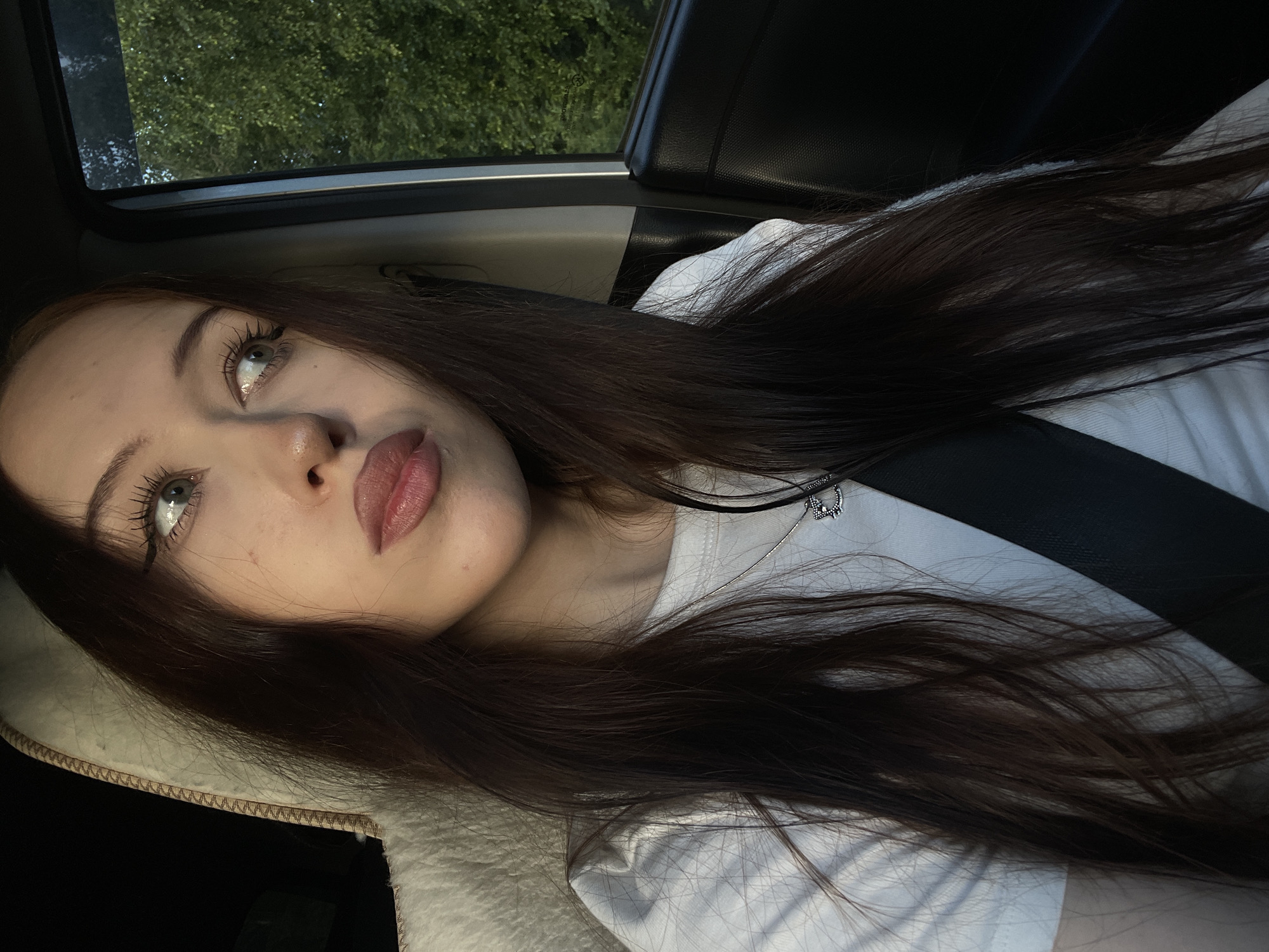 A young woman with long, dark brown hair is captured in a close-up, angled selfie, likely taken inside a vehicle. She is wearing a white t-shirt and a black seatbelt is visible across her chest, suggesting she is traveling. A glimpse of lush green trees through the window indicates an outdoor setting, possibly during the daytime. Her gaze is directed upwards and slightly to the left, with her lips parted. The lighting suggests it could be late afternoon or early evening. A silver necklace with a small pendant is visible on her chest. The overall mood appears relaxed and contemplative. No visible text is present in the image.