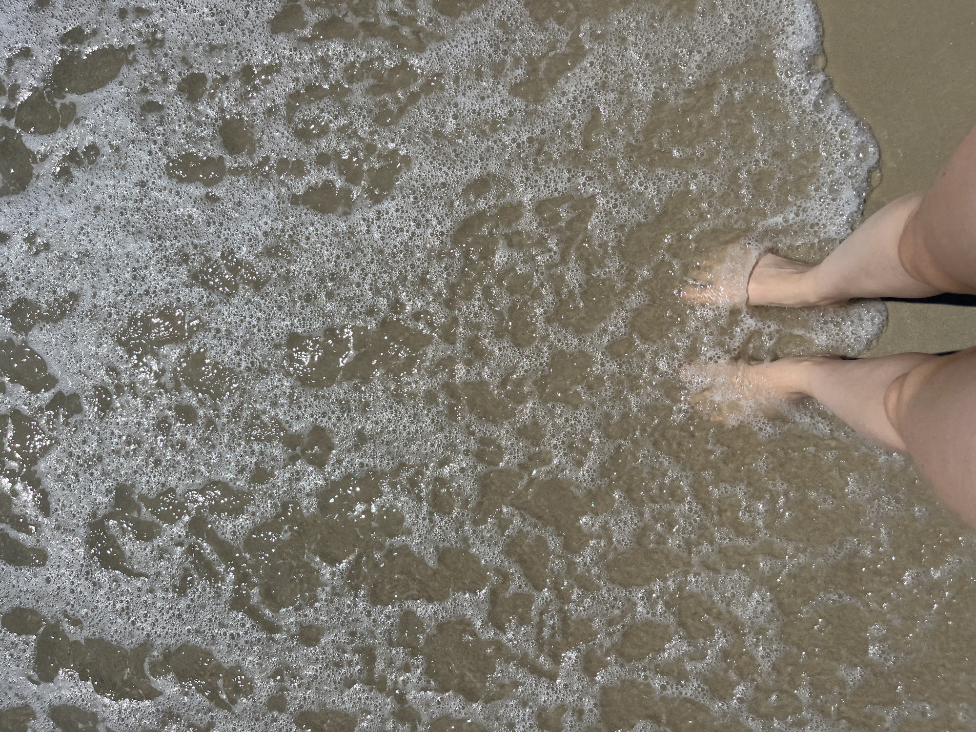 A close-up, top-down view of bare feet standing at the edge of the ocean on a sandy beach. The water is shallow, with foamy waves lapping around the ankles. The sand appears light brown and wet. The lighting suggests it is daytime, likely sunny, with reflections visible on the water's surface.