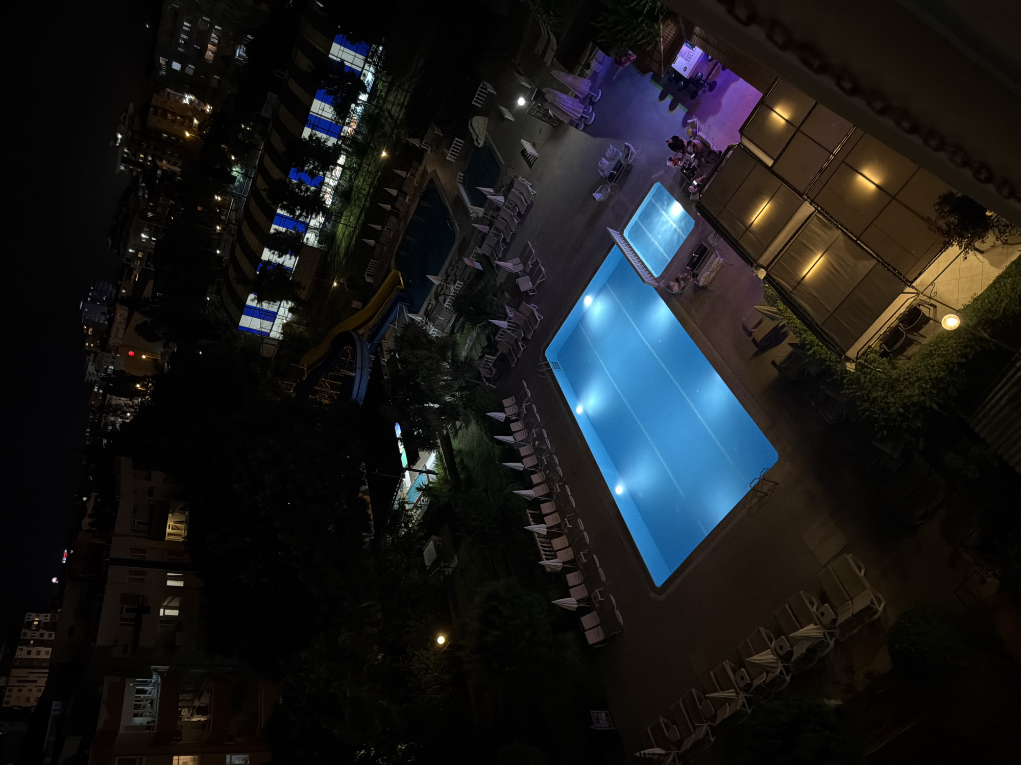 The image shows an outdoor swimming pool area at night. A large, rectangular swimming pool with bright blue lights illuminates the water. Several lounge chairs are arranged around the pool, many with umbrellas. To the left of the main pool, there is a smaller, rectangular pool. A yellow water slide is partially visible. In the background, buildings with illuminated windows are seen, suggesting an urban or resort setting. Some trees and greenery are also present. A few people are visible near a shaded area with tables and chairs, possibly a bar or restaurant, where they appear to be socializing. The scene is lit by the pool lights, overhead lights, and lights from the buildings. The time of day is clearly night. There are no visible signs of specific weather conditions. The overall atmosphere is tranquil and inviting, with the illuminated pool creating a focal point.