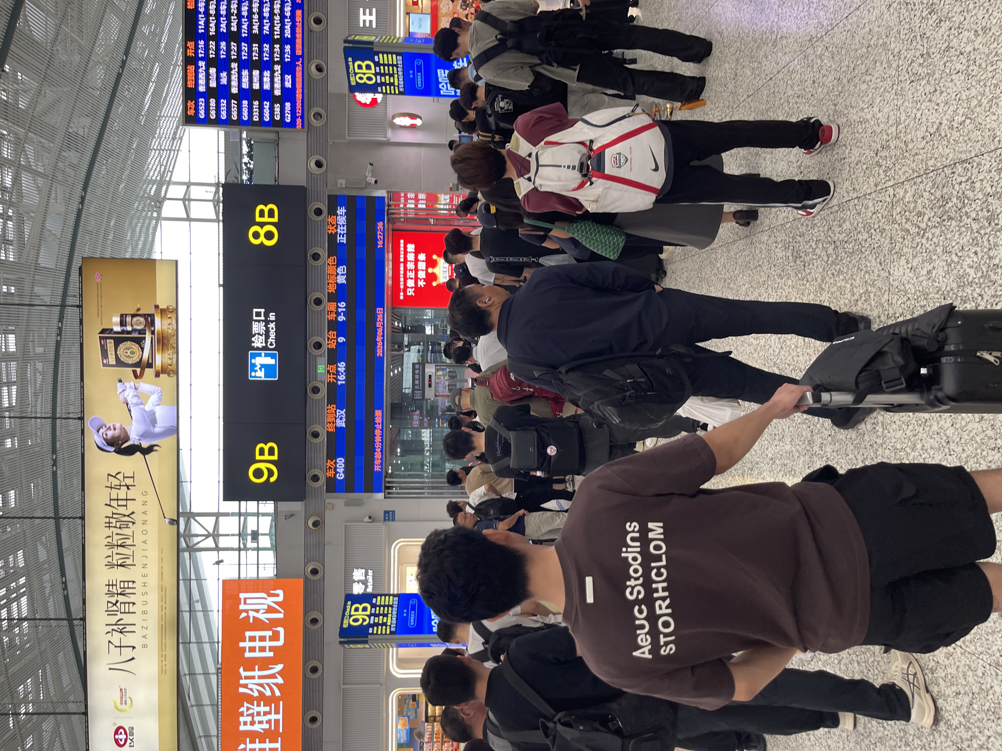 This image captures a busy scene at a train station in Longhua District, China. A crowd of people, dressed in casual attire, are standing or walking, many with luggage. The environment is indoors with a high ceiling and fluorescent lighting. Large digital displays are prominently featured, showing train information such as departure times, destinations, and gate numbers (e.g., "9B", "8B"). One display indicates "Check in" with a pictogram. Advertisements are visible on the walls, including one with Chinese characters and an image of a woman playing golf. The floor is tiled with a speckled pattern. A person in the foreground, facing away from the camera, wears a brown t-shirt with white text reading "Aeuc Stodins STORHCLOM" and black shorts, holding a rolling suitcase. Another person nearby is wearing a white and red backpack. The overall atmosphere suggests transit activity and waiting for trains.