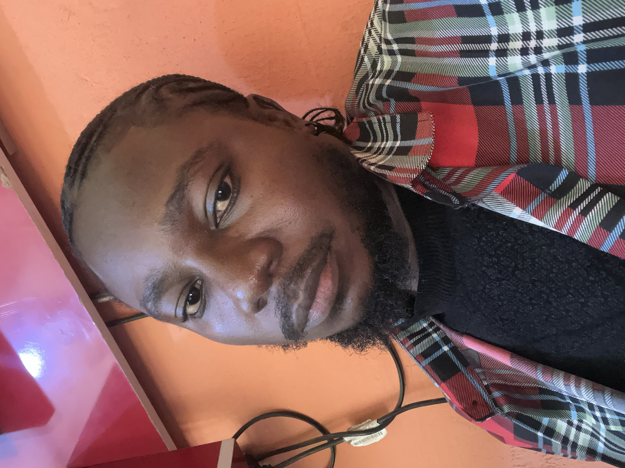 In a medium close-up shot, a young Black man with braided hair and a beard is pictured indoors against an orange wall. He is looking directly at the camera. He is wearing a black turtleneck sweater underneath an open, multi-colored plaid shirt featuring red, black, green, blue, and white patterns. A dark earring is visible on his left ear. To the left of the subject, black electrical cords are bundled together. Above and behind him, a bright red object, possibly a piece of furniture or equipment, is partially visible. The lighting suggests an indoor setting, possibly a room in Jos, Nigeria, given the visual cues of the environment and the subject's appearance.