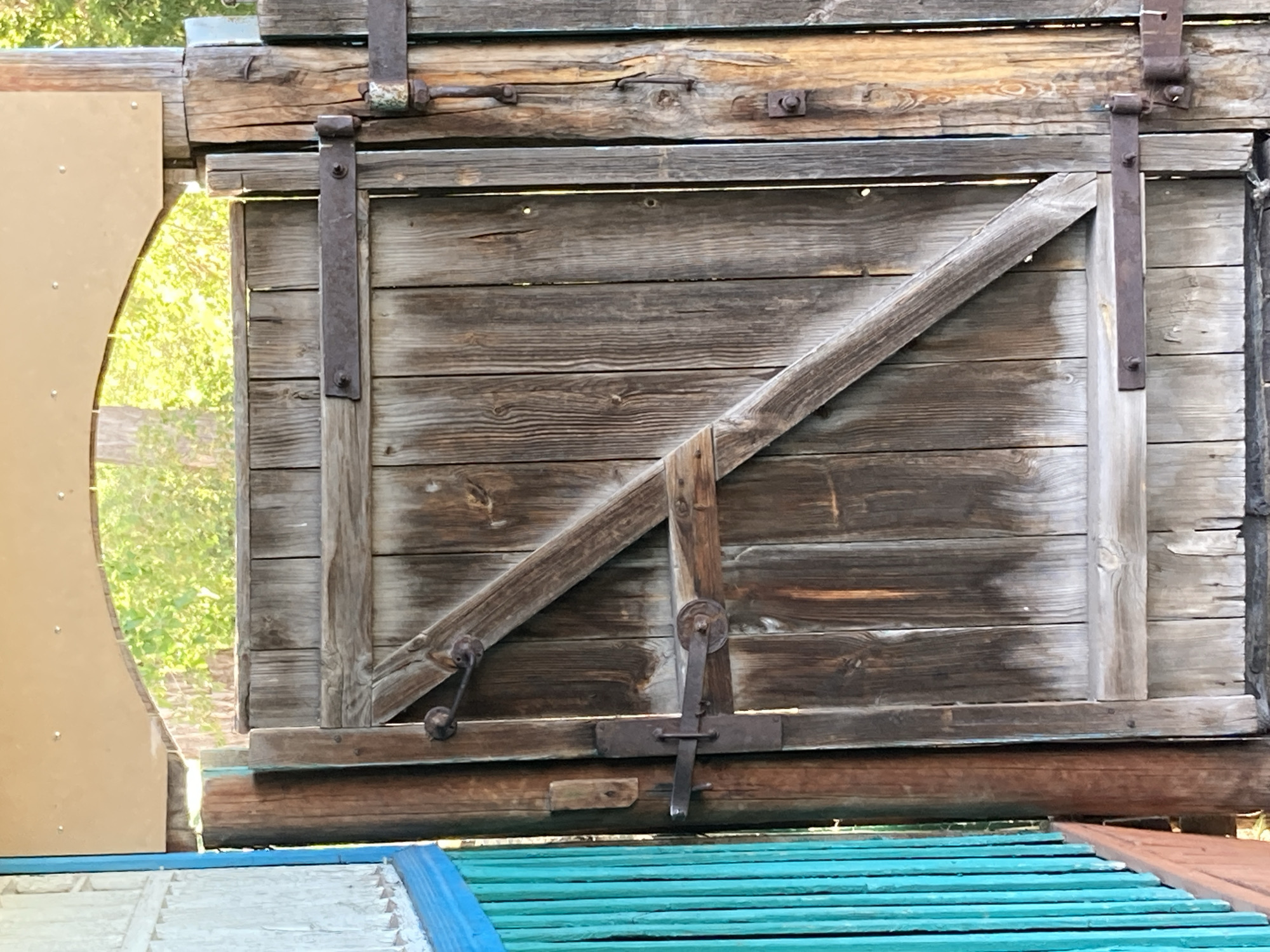The image shows a weathered wooden door with metal hinges and latches. The door is part of an old structure, possibly a barn or shed, made of rough-hewn wooden planks. A diagonal wooden brace reinforces the door. To the left, a curved beige panel with visible rivets suggests a modern addition or covering. Through an opening in the door, lush green foliage and dappled sunlight indicate an outdoor setting during the daytime, likely with pleasant weather. Below the door, sections of brightly colored wooden planks, painted in blue and green, are visible, hinting at a playful or decorative element. The scene evokes a sense of rustic charm and history. No people are visible in the image, and there is no visible text.
