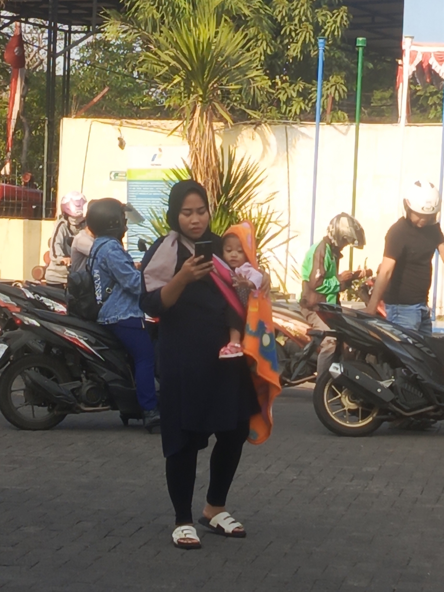 In Surabaya, Indonesia, a woman in a dark blue dress and black headscarf is holding a baby wrapped in an orange and blue blanket, looking at her phone. The baby is wearing pink sandals. The woman is standing on a paved surface with motorcyclists in the background. One motorcyclist, wearing a green jacket and a helmet, is also looking at a phone. Another motorcyclist, in a black shirt and helmet, is standing beside a motorcycle. Two other motorcycles are parked, and several people on motorcycles are visible in the distance, one wearing a pink helmet. Trees and a yellow building with signage are in the background.
