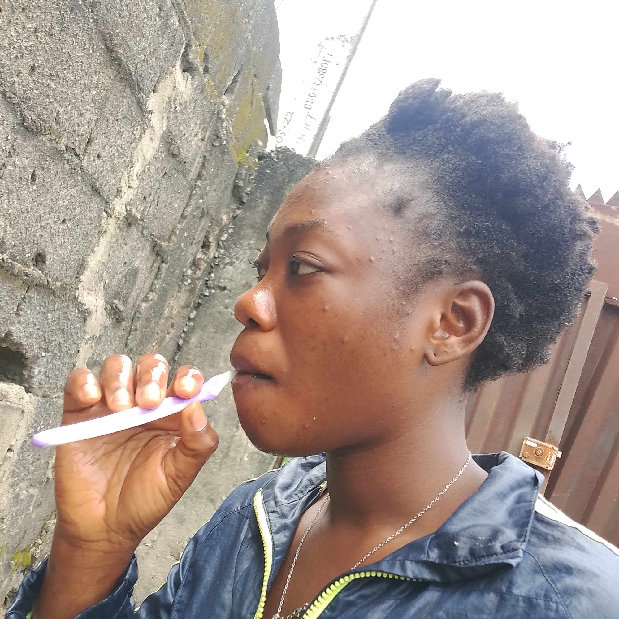 A medium shot captures a young Black woman in profile, brushing her teeth with a purple toothbrush against a rough, textured concrete wall. She is wearing a dark blue jacket with a neon green zipper. The background includes a brown corrugated metal fence with a small gold lock. The woman's hair is dark and styled in a short, textured cut. She has light brown skin with visible pores and a few blemishes on her face. The lighting suggests an overcast day. The scene appears to be outdoors in a city environment, potentially in Ado, Nigeria, given the context.