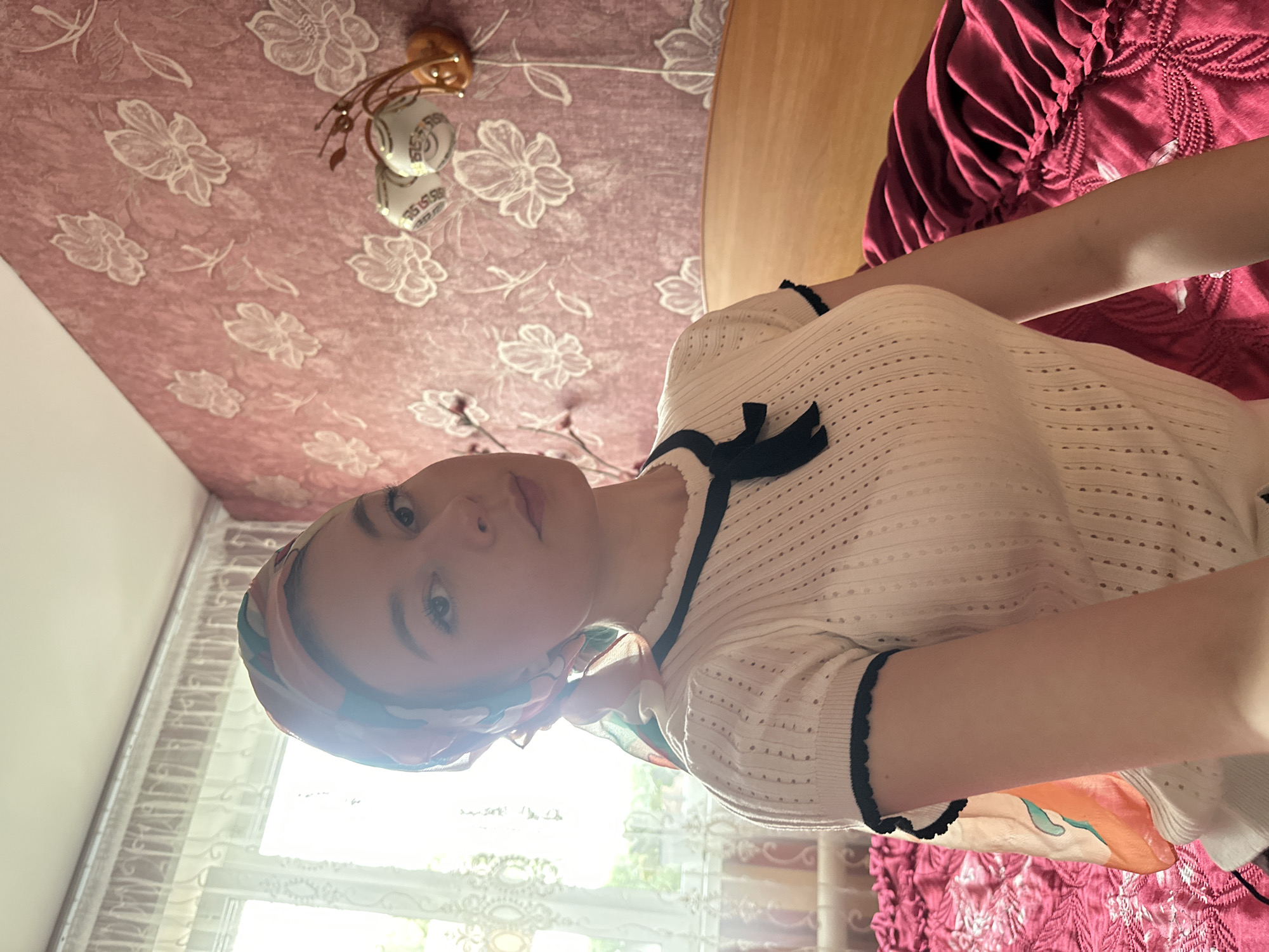 A woman wearing a headscarf and a white, horizontally striped shirt with black trim is in a room with floral wallpaper and curtains, possibly in a private residence. The orientation of the photograph is such that the ceiling is on the left and a window on the right. The scene is brightly lit by natural light coming through the window. The city of Коноваловка, Russia, is not explicitly mentioned or evidenced in the image.