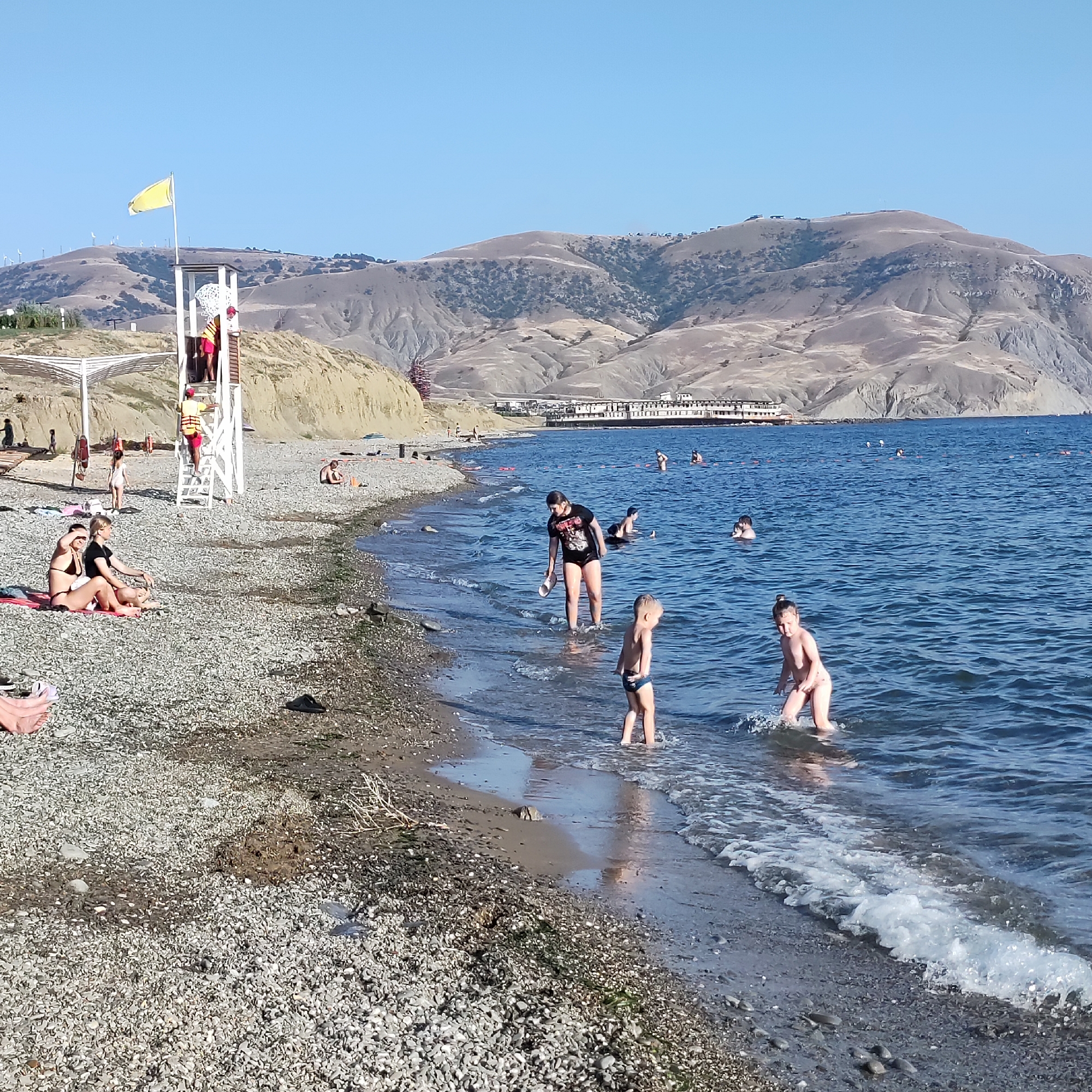 The image shows a crowded beach on a sunny day. The beach is covered with pebbles. In the foreground, two women are sitting on a towel, wearing bikinis. To their right, a child is standing in the shallow water. In the midground, several people are in the sea, some swimming and others wading. There is a lifeguard tower on the left side of the beach with two lifeguards. In the background, there are hills with some buildings on them, and the sea stretches out to the horizon. The sky is clear and blue.