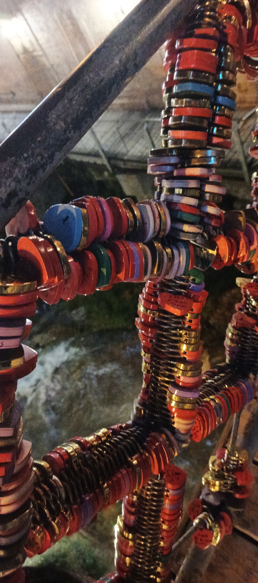 The image shows a section of a metal railing densely adorned with colorful, circular love locks. These locks, primarily red with gold accents, are attached to the railing, creating a layered effect. A single blue love lock is also visible. The background reveals a flowing body of water, likely a river or canal, suggesting the railing is part of a bridge. The scene is located in Tbilisi, Georgia, where such love lock installations are common tourist attractions on bridges over the Kura River.